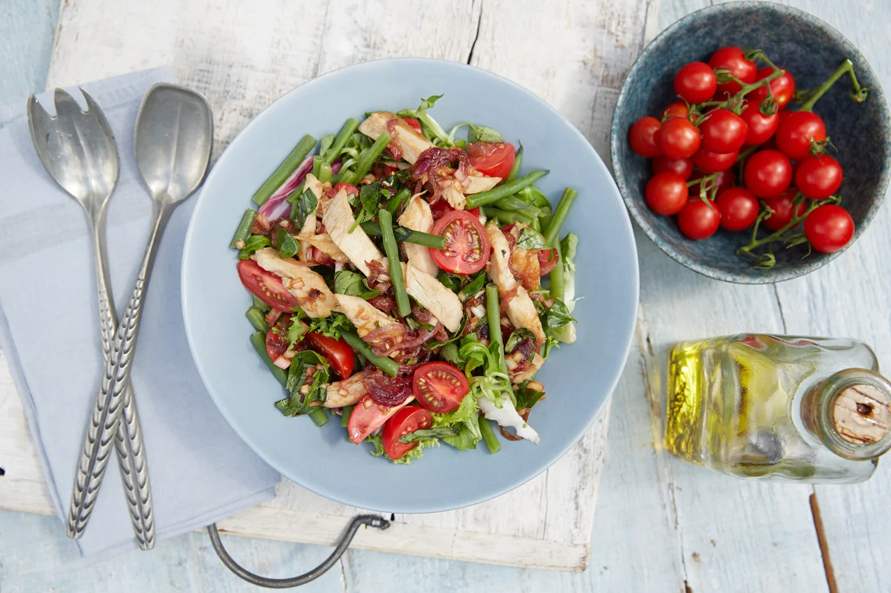 A light blue plate of garden salad with cherry tomatoes, green beans, and grilled chicken on a white wooden table, with a bowl of cherry tomatoes, a bottle of olive oil, and salad utensils nearby.