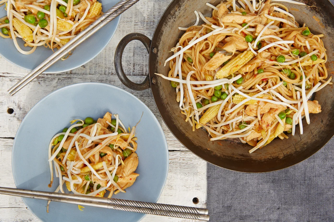 Stir fried noodle dish with chicken, bean sprouts, peas, and baby corn served on a large wok and two blue plates with chopsticks.