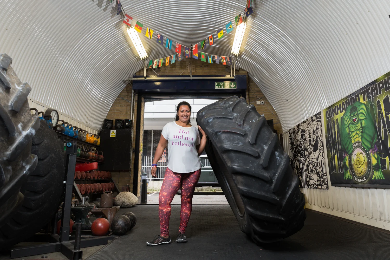 A woman smiling and touching a large black tire in a gym or training area with colorful banners hanging from the ceiling, weightlifting equipment on the side, and a motivational poster on the wall.