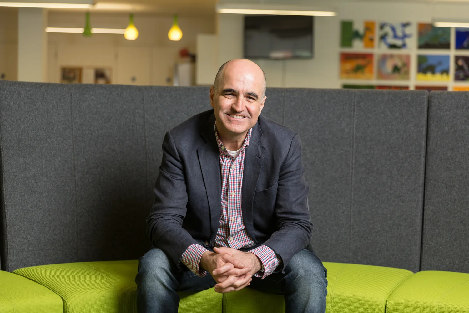 A smiling man with a shaved head, wearing a black blazer over a red and white checkered shirt, sitting on a green couch in a modern office space with colorful wall art and hanging pendant lights.