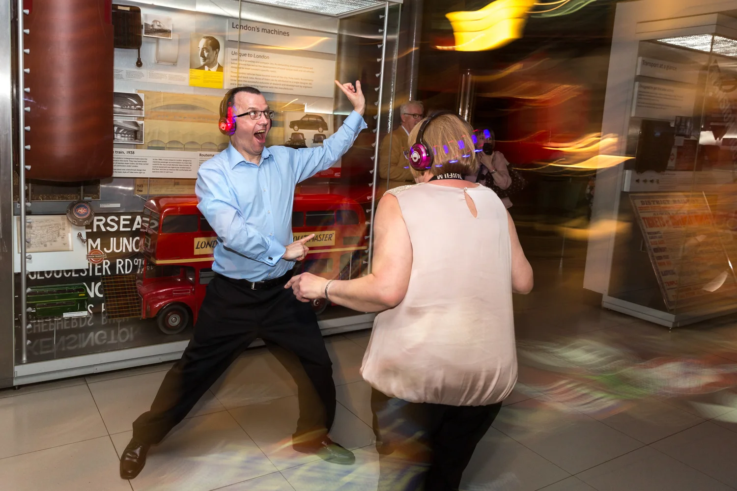 Two people wearing headphones dance and enjoy music in front of a display about London's transportation history at a museum.