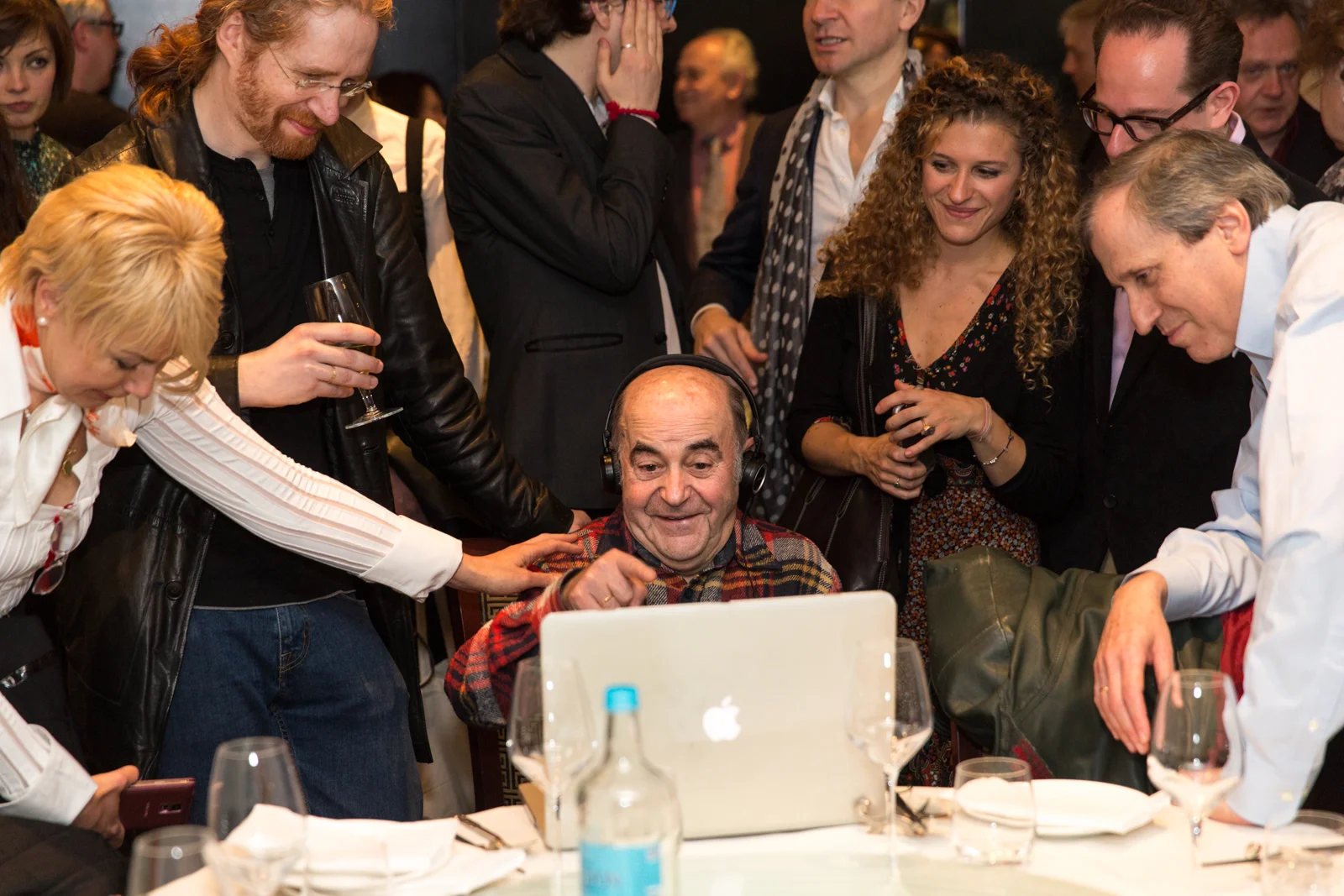 A group of people gathered around a man wearing headphones and sitting in front of a laptop at a social event. The group appears to be enjoying a moment of entertainment or discussion.