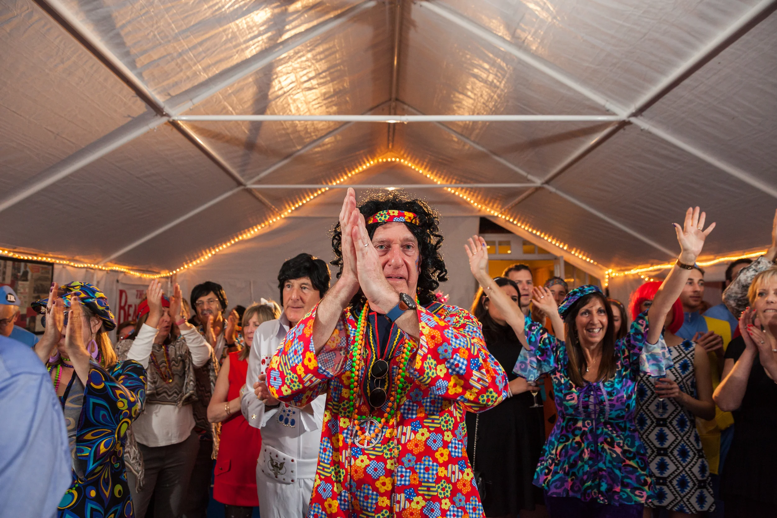 People dressed in colorful 1960s hippie costumes dancing and having fun at a party under a decorated tent.