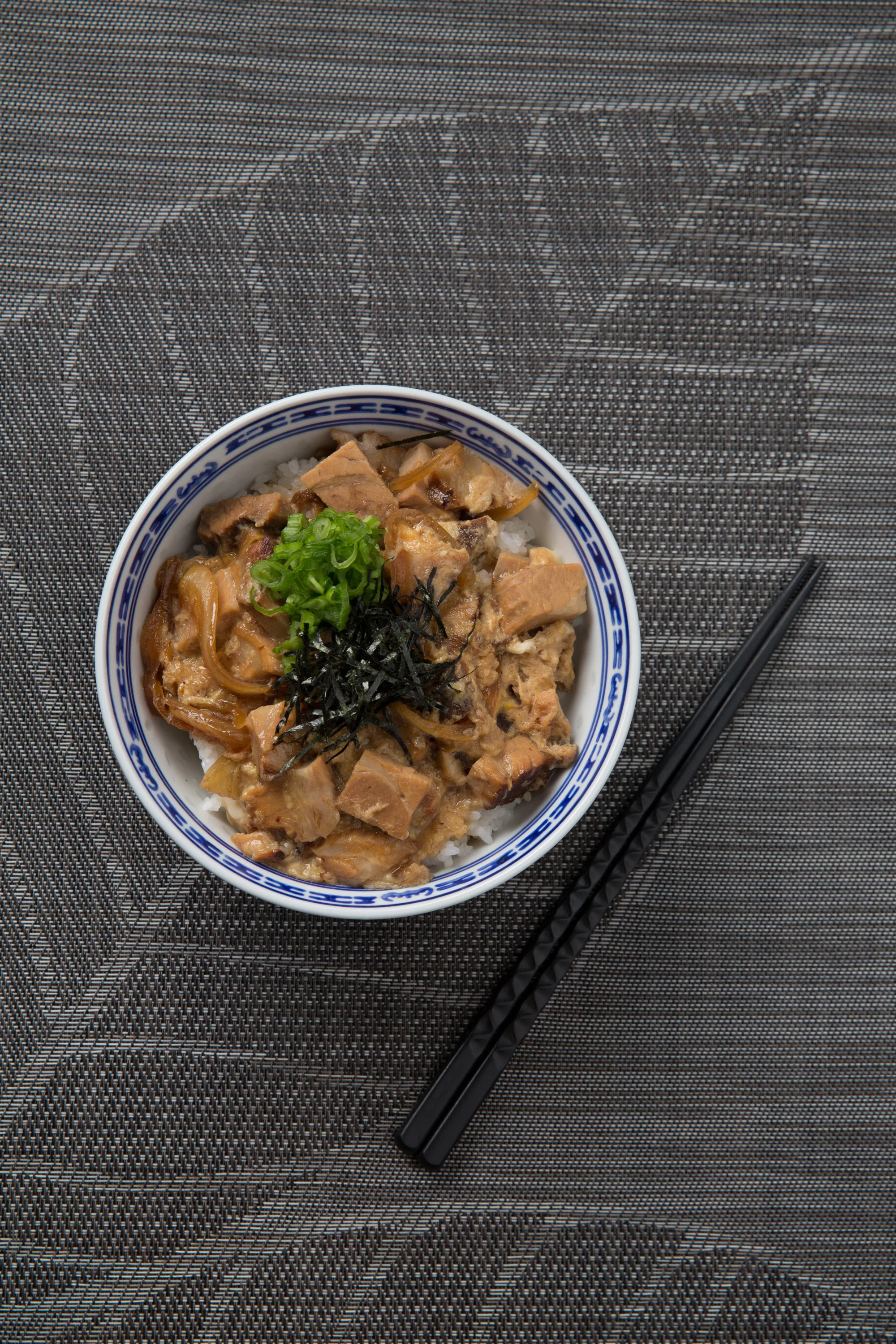 Bowl of rice topped with chicken, onions, green onions, and shredded seaweed, on a woven placemat with black chopsticks.