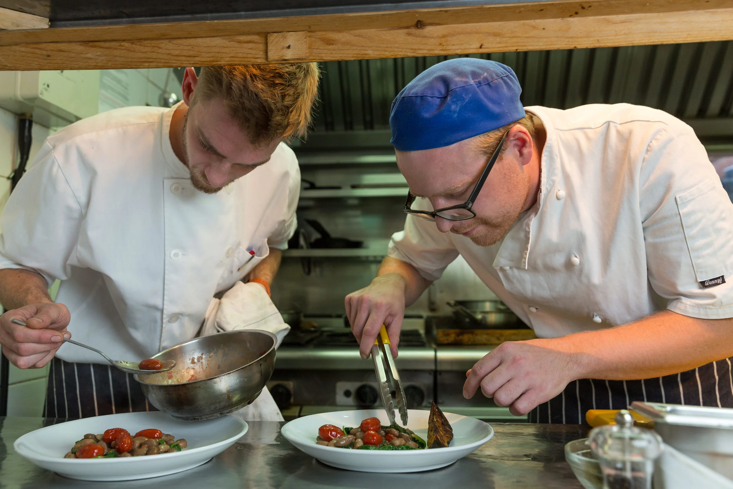 Two male chefs in white uniforms preparing plated dishes in a commercial kitchen.
