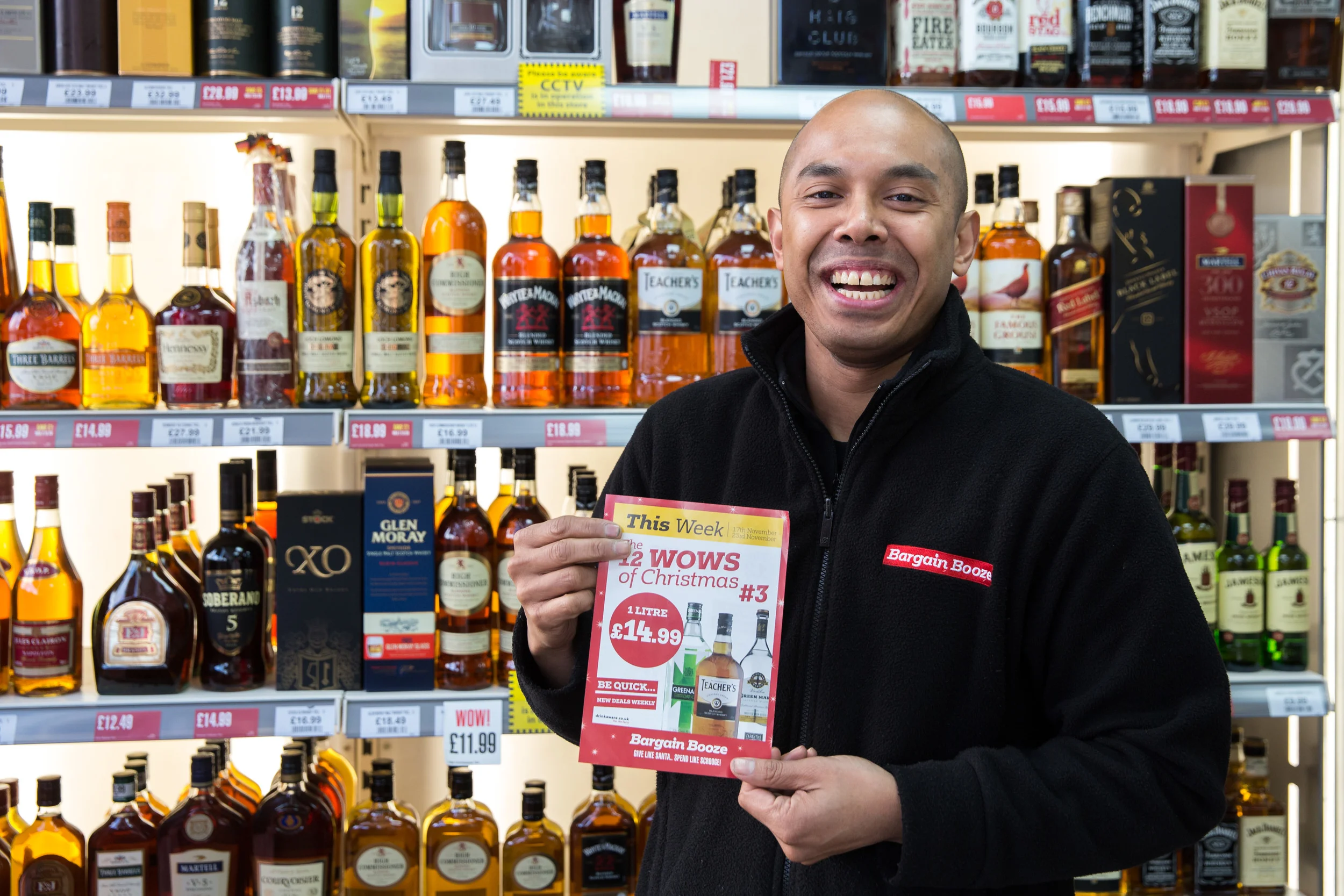 A smiling man in a black jacket in a liquor store holding a flyer advertising a Christmas liquor sale, with shelves of various bottles of whiskey and other spirits in the background.