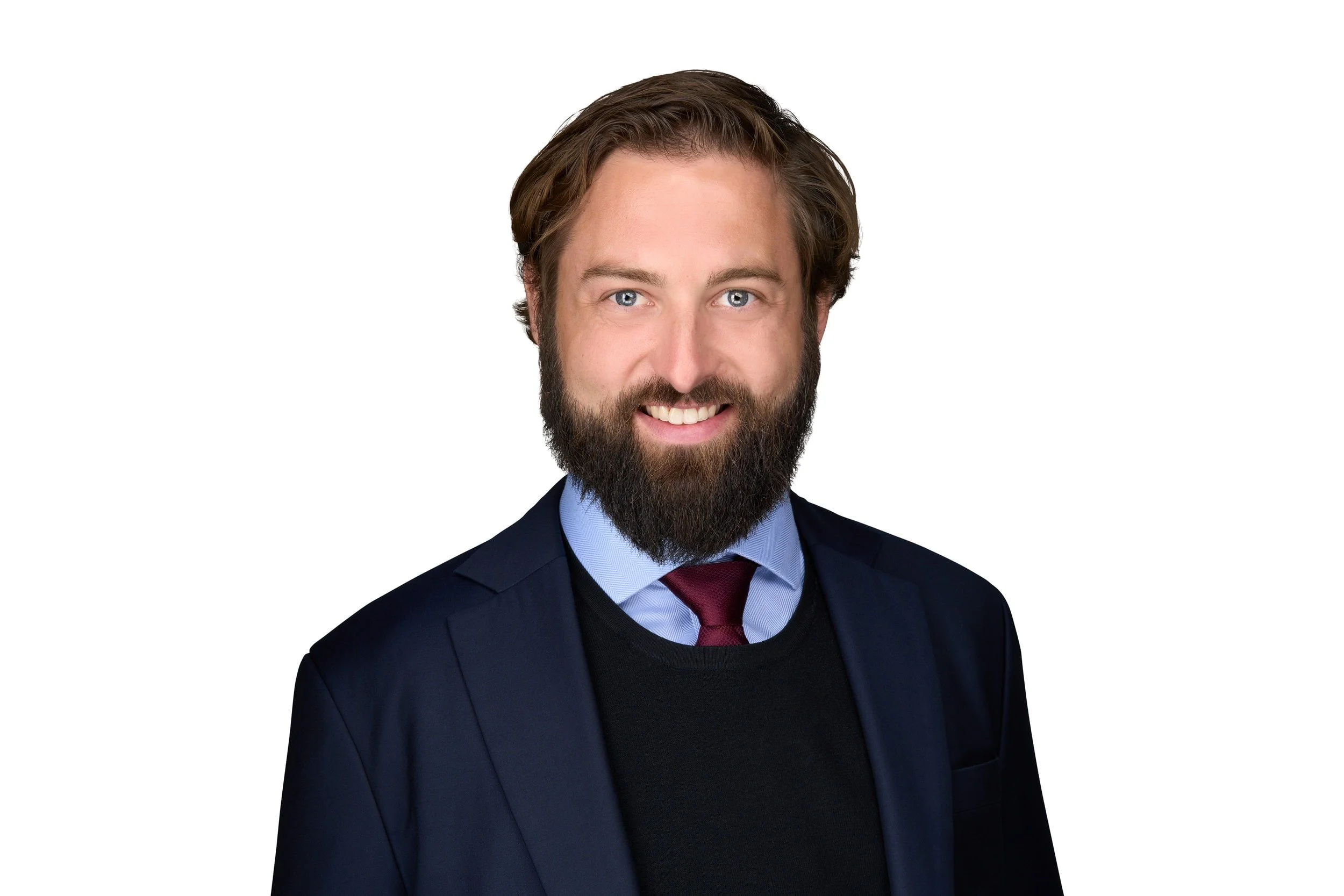 Portrait of a man with a beard wearing a dark blazer, light blue shirt, black sweater, and maroon tie, smiling against a white background.