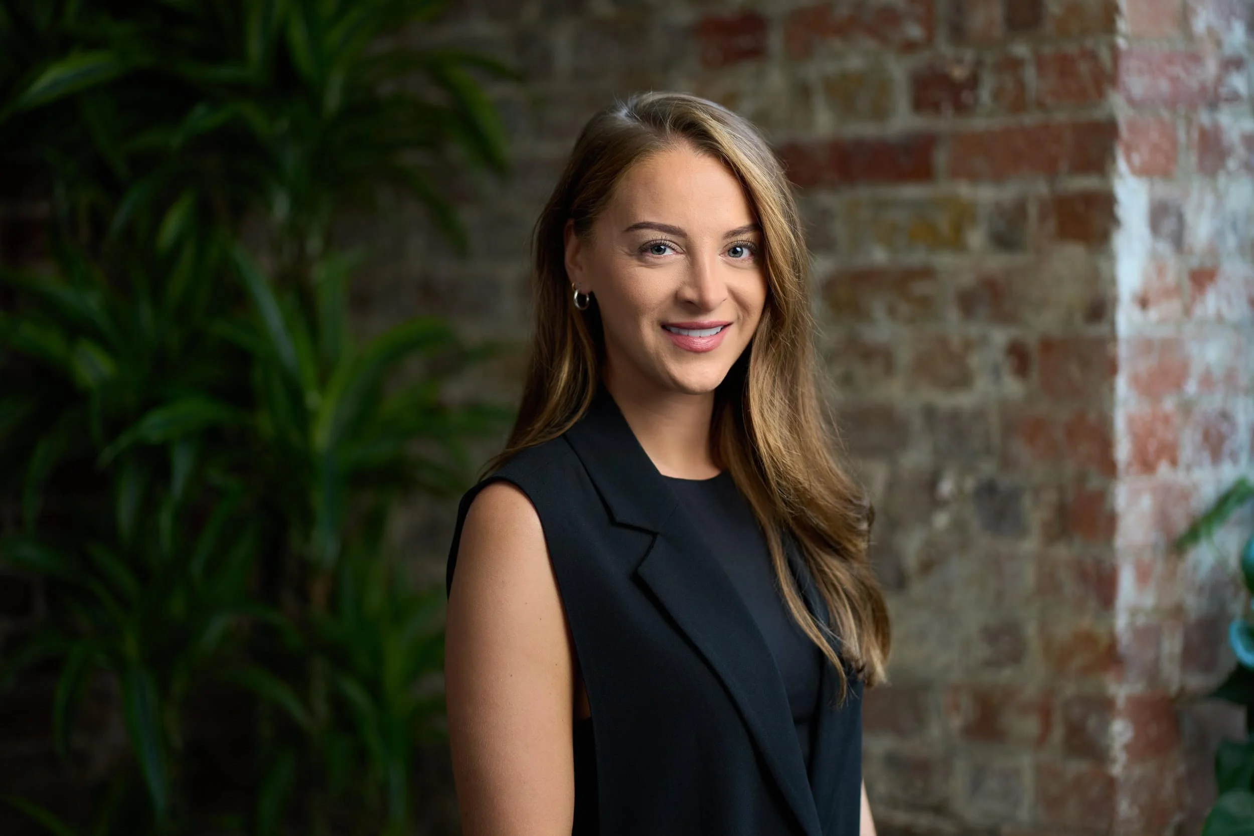 A woman with long brown hair smiling, wearing a black sleeveless blazer, standing indoors against a brick wall and green plants.
