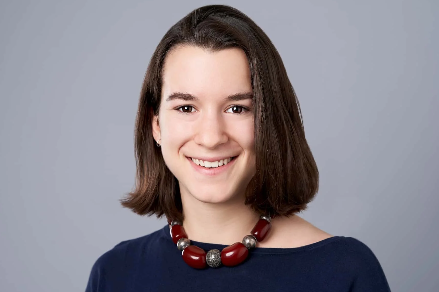 Young woman with shoulder-length brown hair wearing a navy blue top and a red and silver beaded necklace, smiling against a plain gray background.
