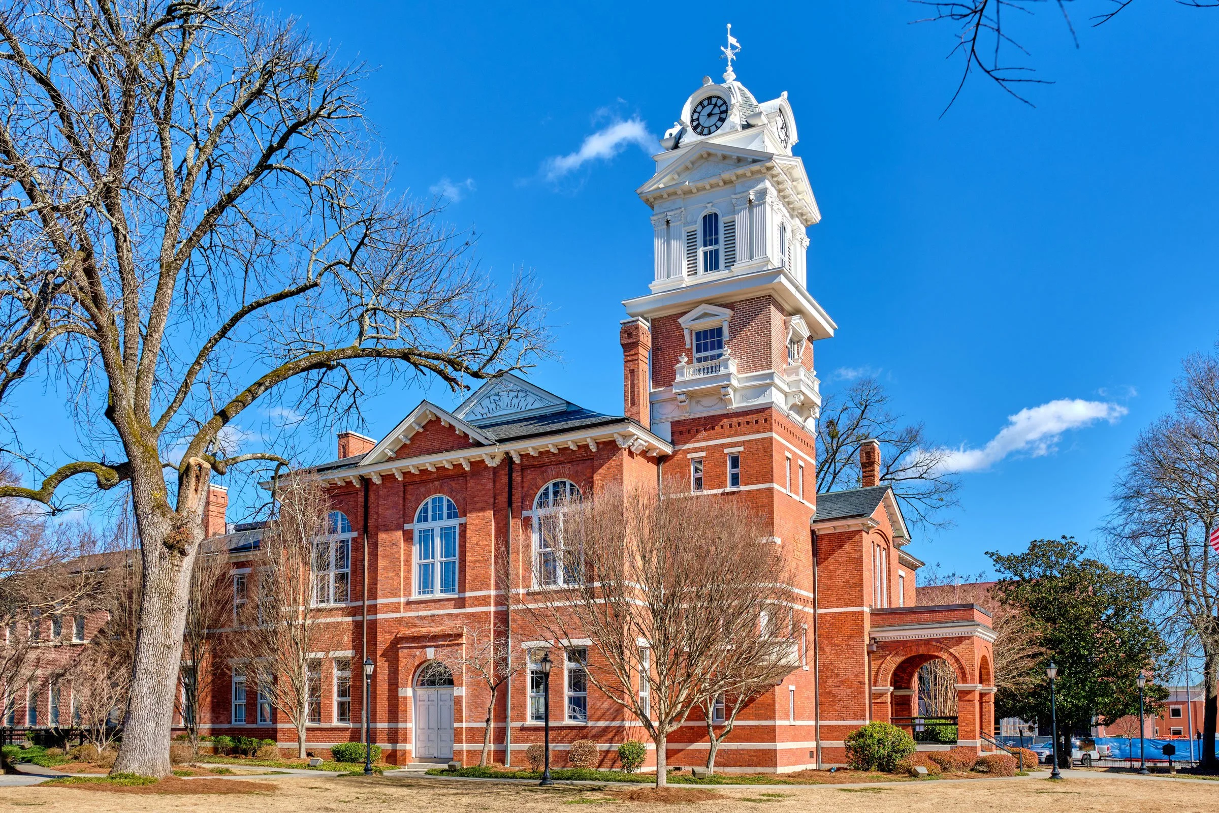 A historic red brick building with a white clock tower under a bright blue sky. Bare trees are in the foreground, indicating a winter or early spring day.