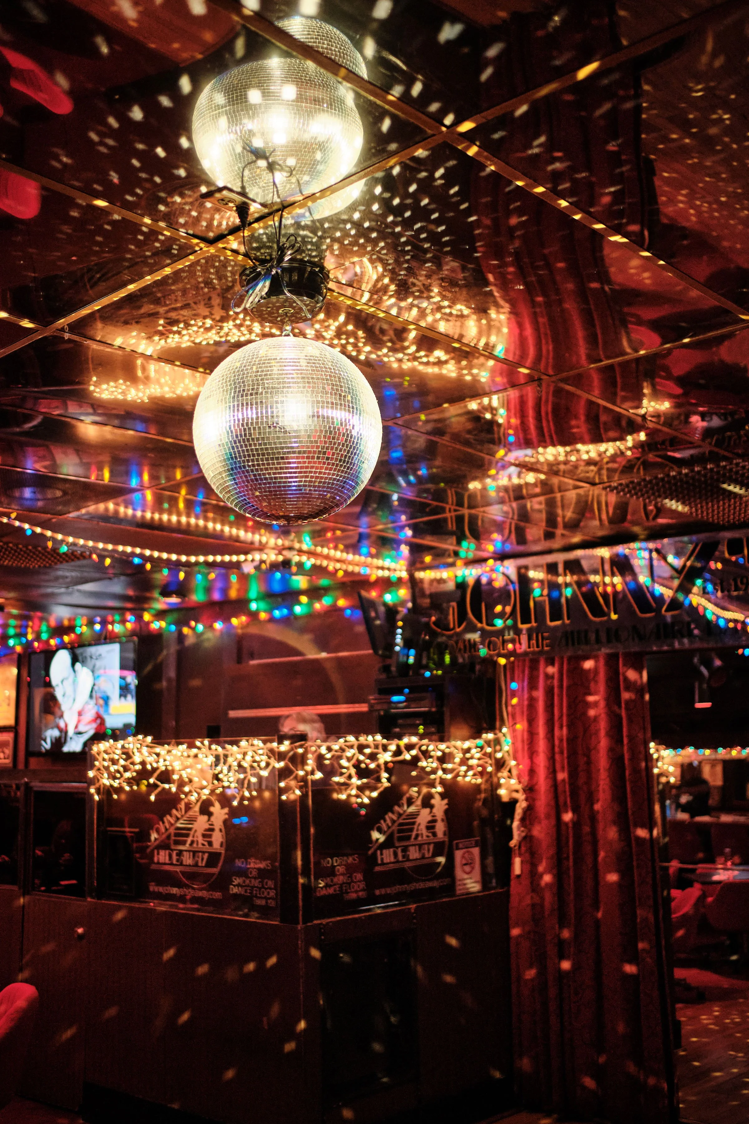 Dance floor area illuminated by a disco ball reflecting light, with colorful string lights and red curtains in the background, indicating a nightclub or party venue.