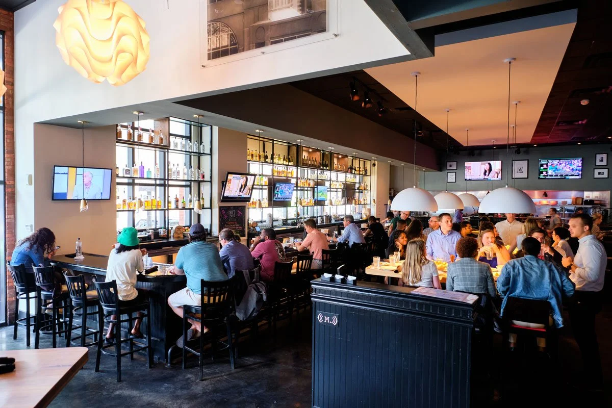 People dining inside a modern bar or restaurant, with shelves of bottles behind the bar, multiple flat-screen TVs on the walls, and hanging pendant lights over the tables.