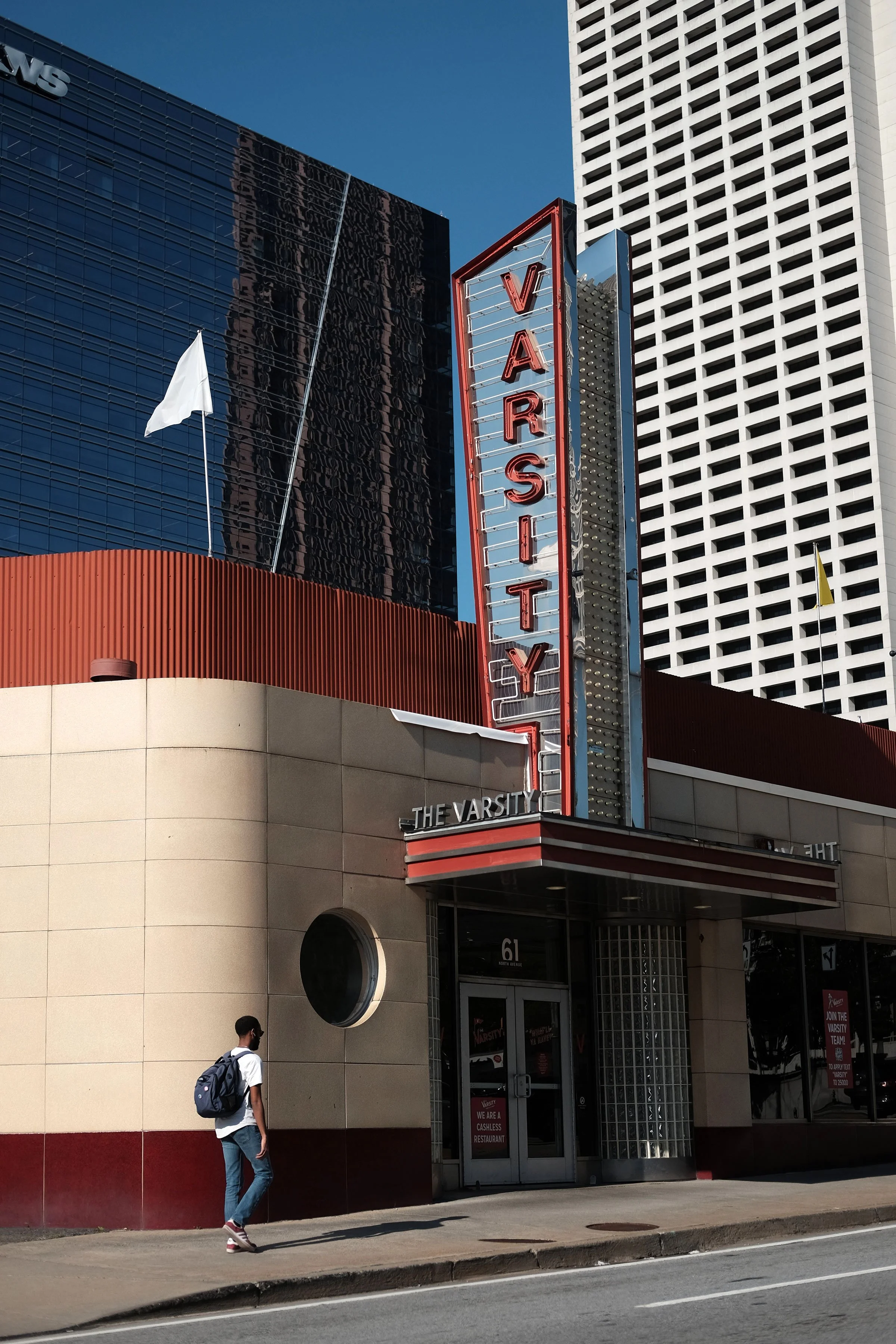 A person wearing a backpack and face mask walking past a building with a sign that reads 'VARSITY'. The building has a modern facade with a combination of tiled and glass windows, and the sky is clear.