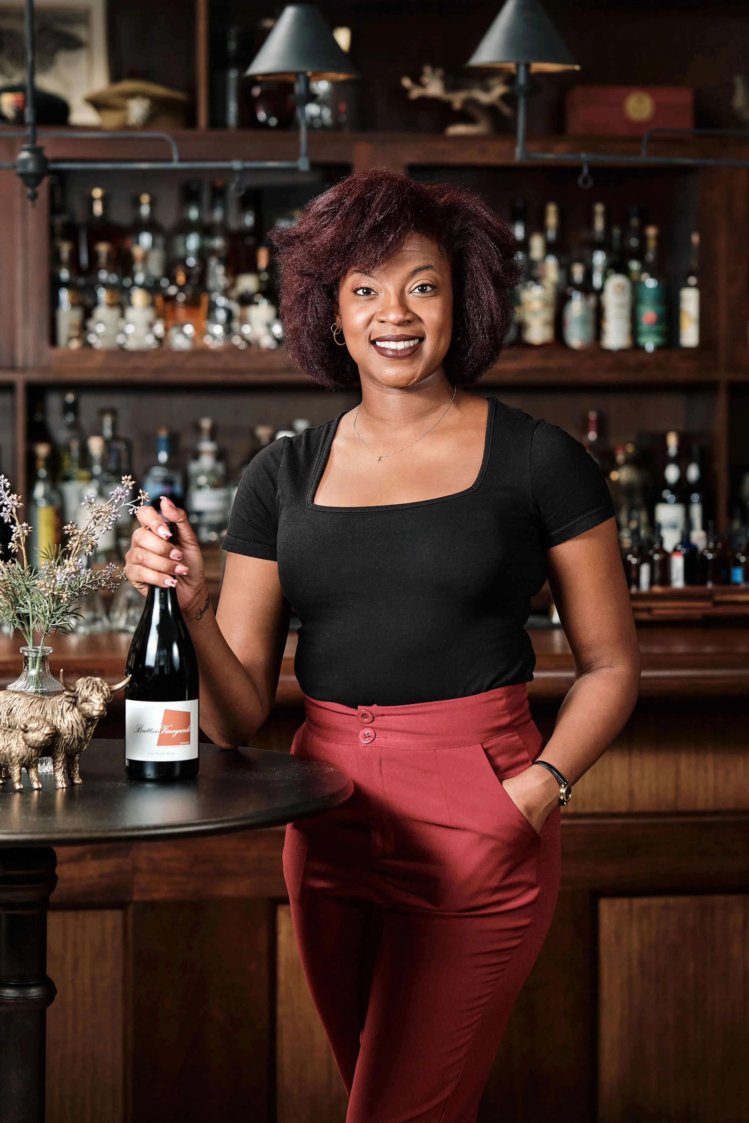 A woman with natural curly hair, wearing a black top and red high-waisted pants, standing in a bar with a dark wooden shelf filled with liquor bottles in the background, holding a bottle of wine.