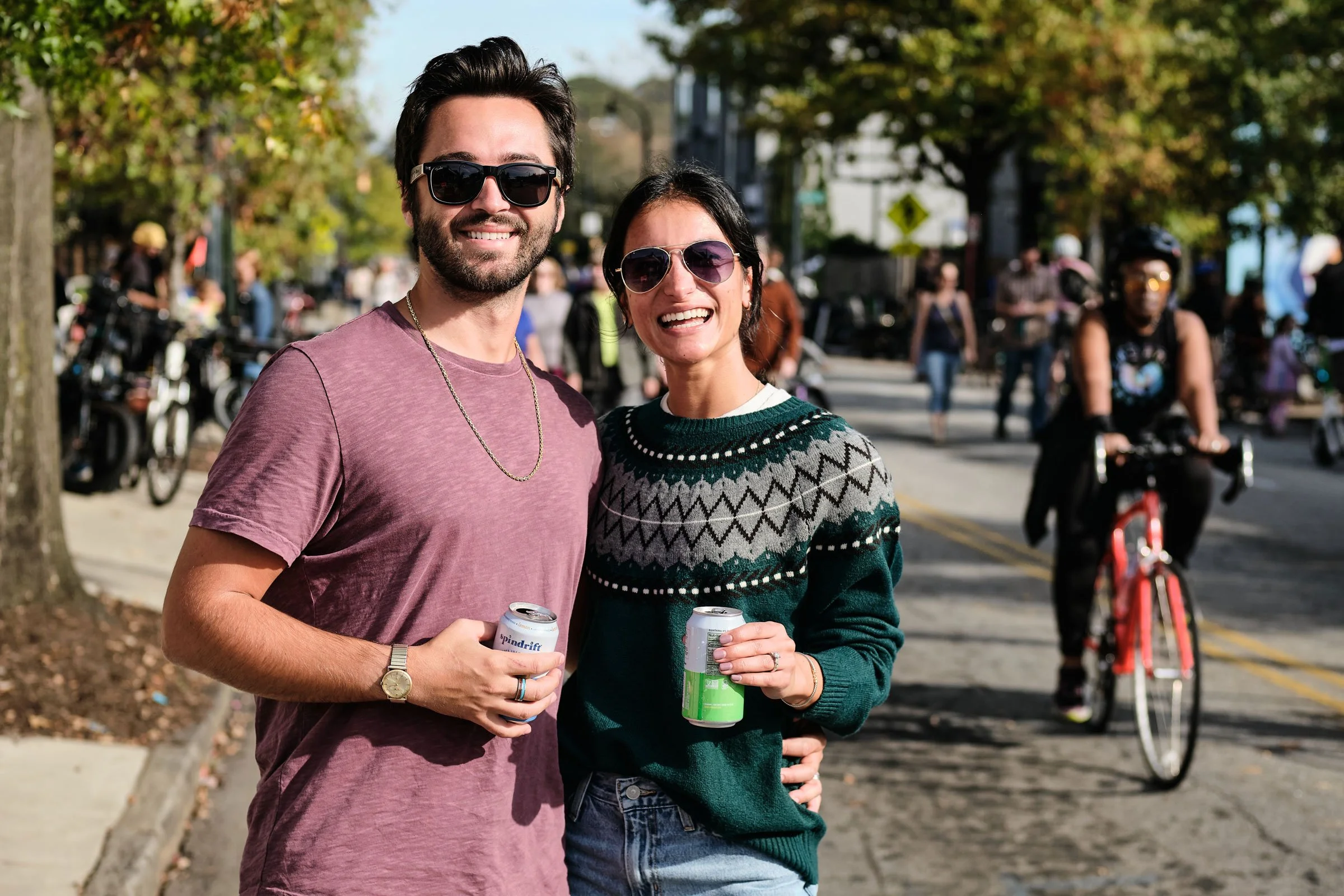 A smiling man and woman standing outdoors on a sunny day, holding canned beverages, with trees, people, and bicycles in the background.