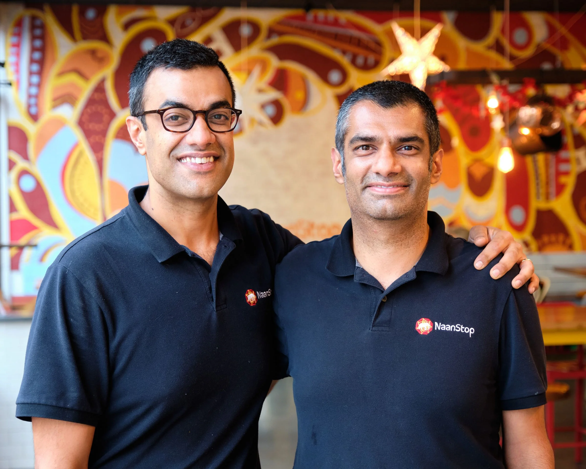 Two smiling men wearing black shirts with a restaurant or café decor background, including a colorful mural and bright star-shaped light in the background.