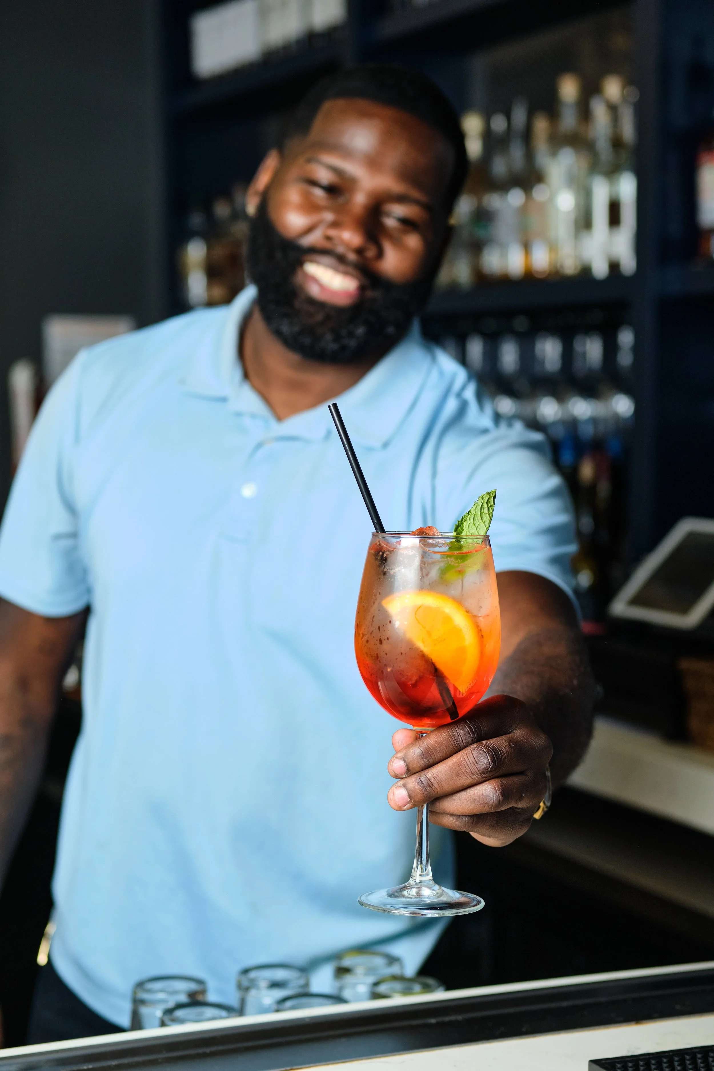 A bartender serving a colorful cocktail with orange, lemon slices, mint leaf, and a straw in a wine glass, behind a bar with shelves of bottles.