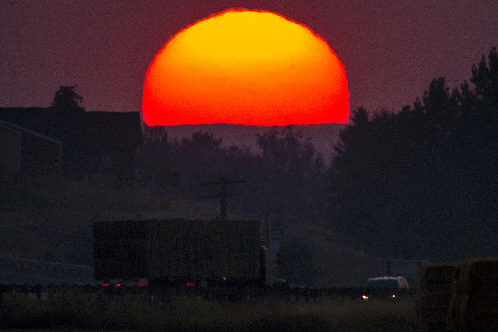 A semi truck hauling bales of hay slows down as it enters Colton, Wash., while the sun begins to set below the hazy horizon on Tuesday, Aug. 7, 2018.