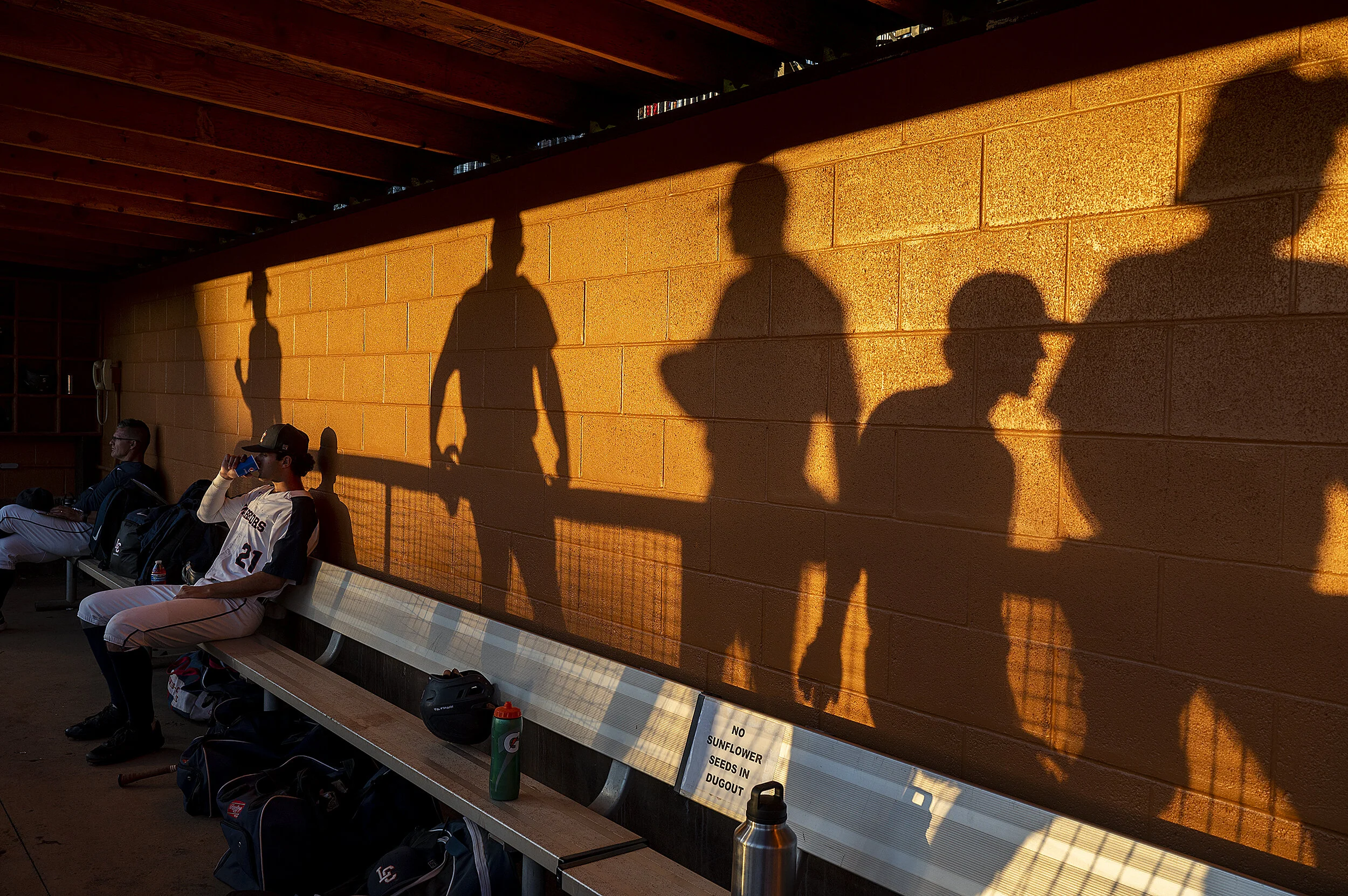  Lewis-Clark State head coach Jake Taylor (left) and Sam Linscott sit in the dugout as shadows of players walking along the outside of the dugout are seen in the bricks behind the bench as the sun sets prior to the Warriors’ opening round game agains