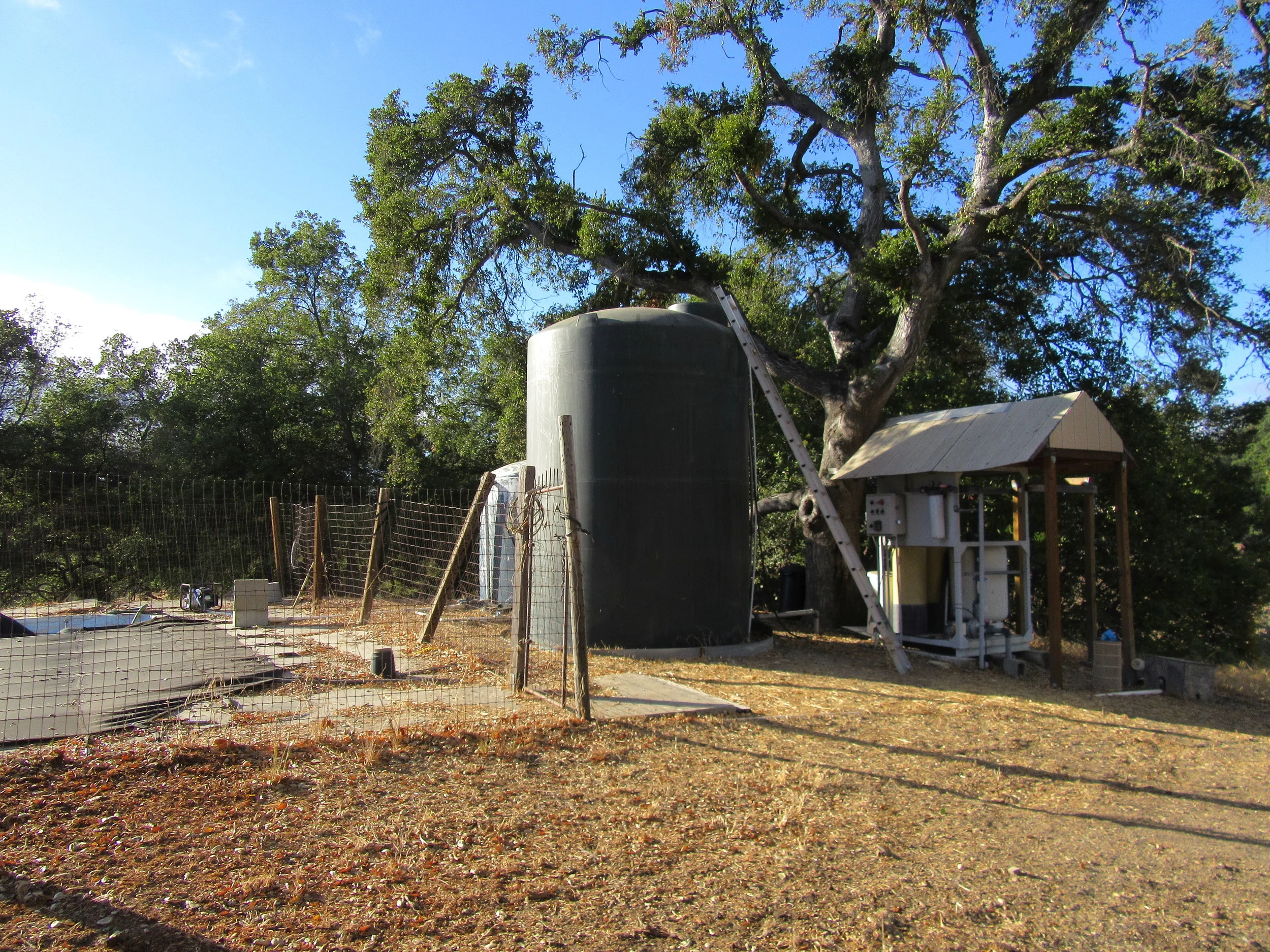 WATER TREATMENT SYSTEM - The former swimming pool, which acts as a reservoir for the water pumped from the well, is to the left enclosed by fencing. The large holding tank in front is for treated water. The smaller holding tank in back is for untreated agricultural water. The water treatment system is to the right. (Photo: Sam Ervin, 2016)