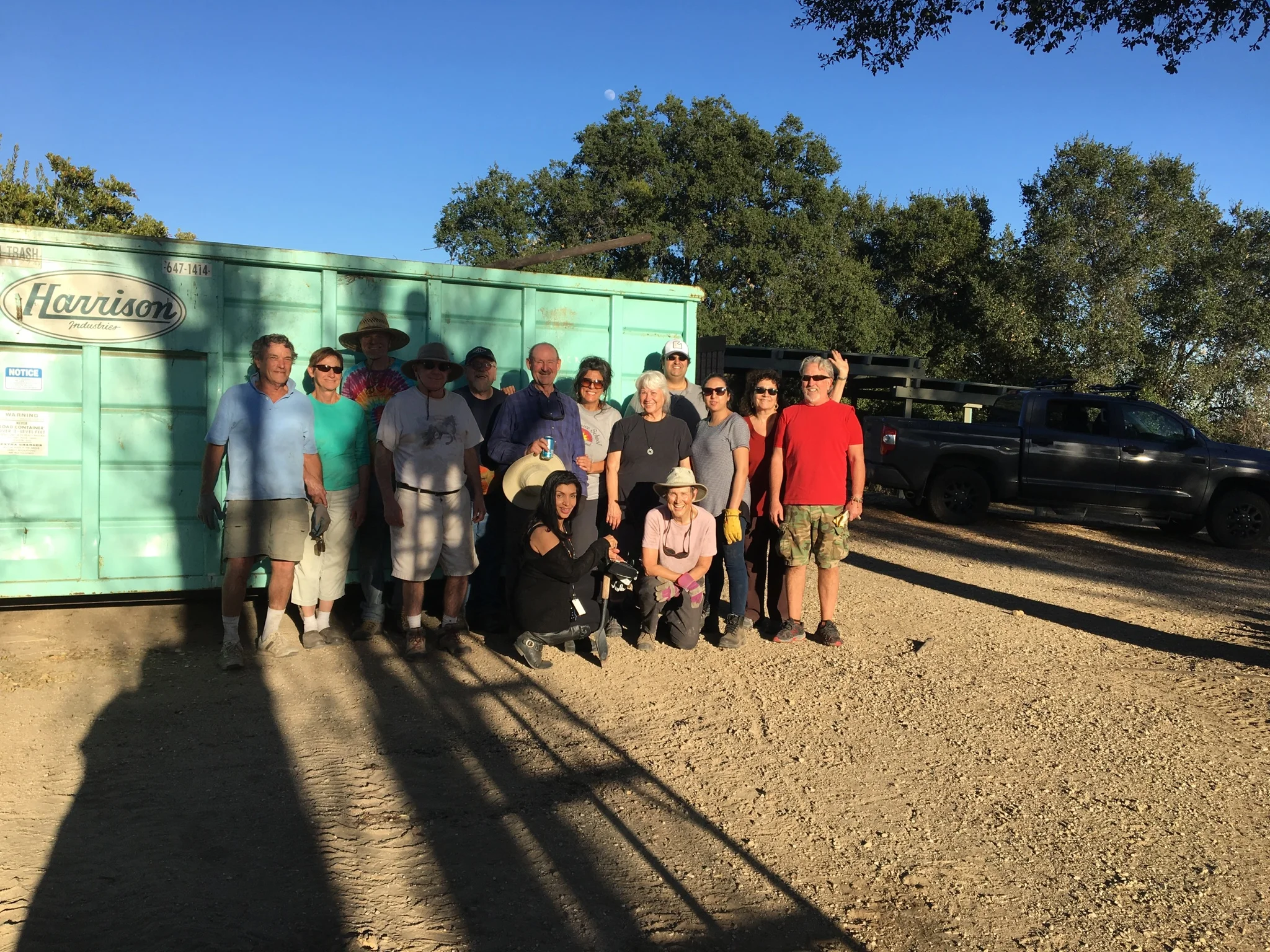 AT DAY'S END on Fire Cleanup Day (January 28, 2018) by the now-full dumpster. (Left to right) Jim Auster, Merrliee Bliss, James Whedon, James Whitson, Buzz Glasky, Sam L. Ervin, Yolanda Koumidou, Ginger Glasky, Homaya Gandi, Khushnam Crawford, Marta Flores, Satya Keyes. (Kneeling) Sarah Larsen, Margaret Magnus. (In shadow) Bing Heckman. (Photo: Cassandra Bramucci)