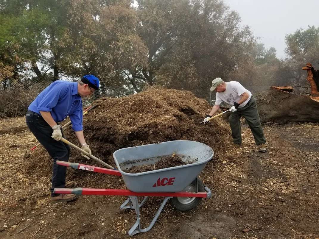 ERIC TURK AND JIM WHITSON load the wheelbarrow with mulch to spread under Baba's Tree at Meher Mount to help hold in the moisture. (Photo: Margaret Magnus, March 10, 2018)