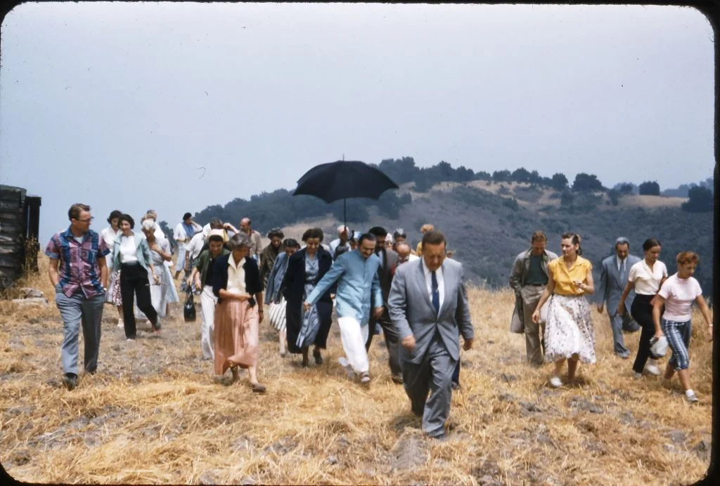 HURRYING TO KEEP UP with Avatar Meher Baba (blue coat) as He tours Meher Mount with co-founder and lifetime caretaker Agnes Baron (light orange skirt) on August 2, 1956. Lud Dimpfl is the man in the suit in front. (Photo: Darwin Shaw, ©Meher Nazar Publications)