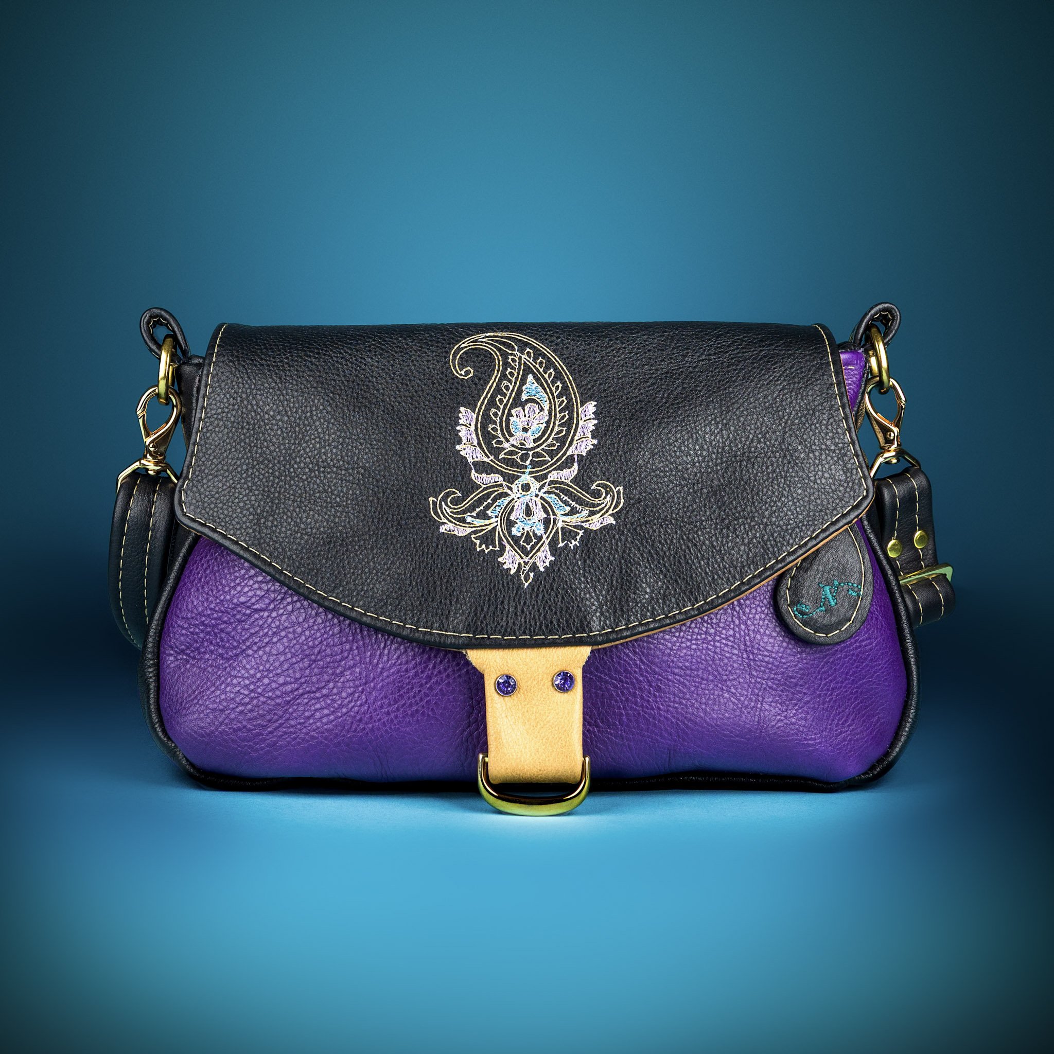 A black purse with gold hardware and embroidered floral designs, purple and yellow accents, against a teal background.