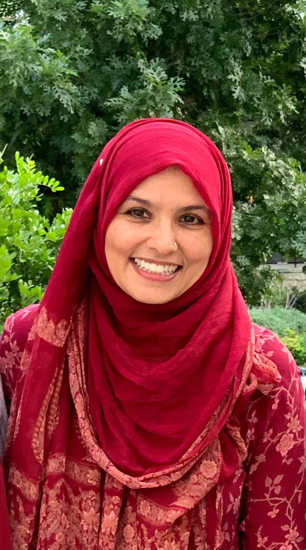 A woman wearing a red hijab and a floral patterned red dress, smiling outdoors with a background of green foliage.