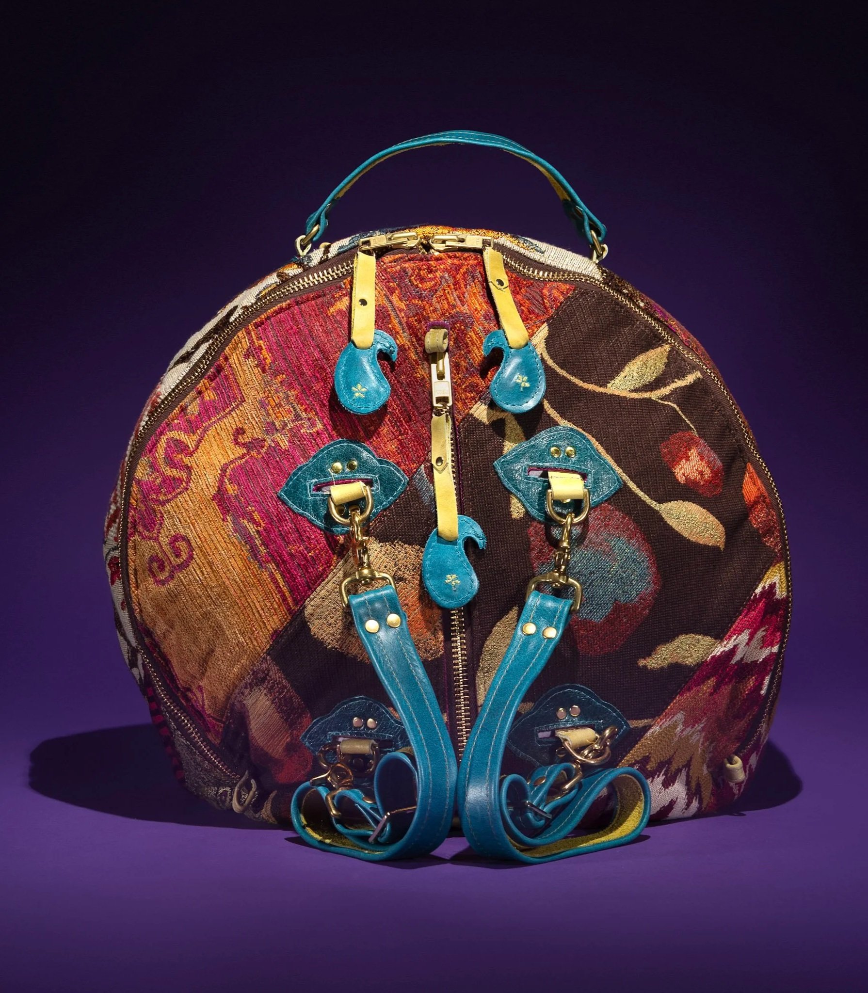 Colorful tapestry backpack with blue leather details, zipper, and chains, set against a dark purple background.