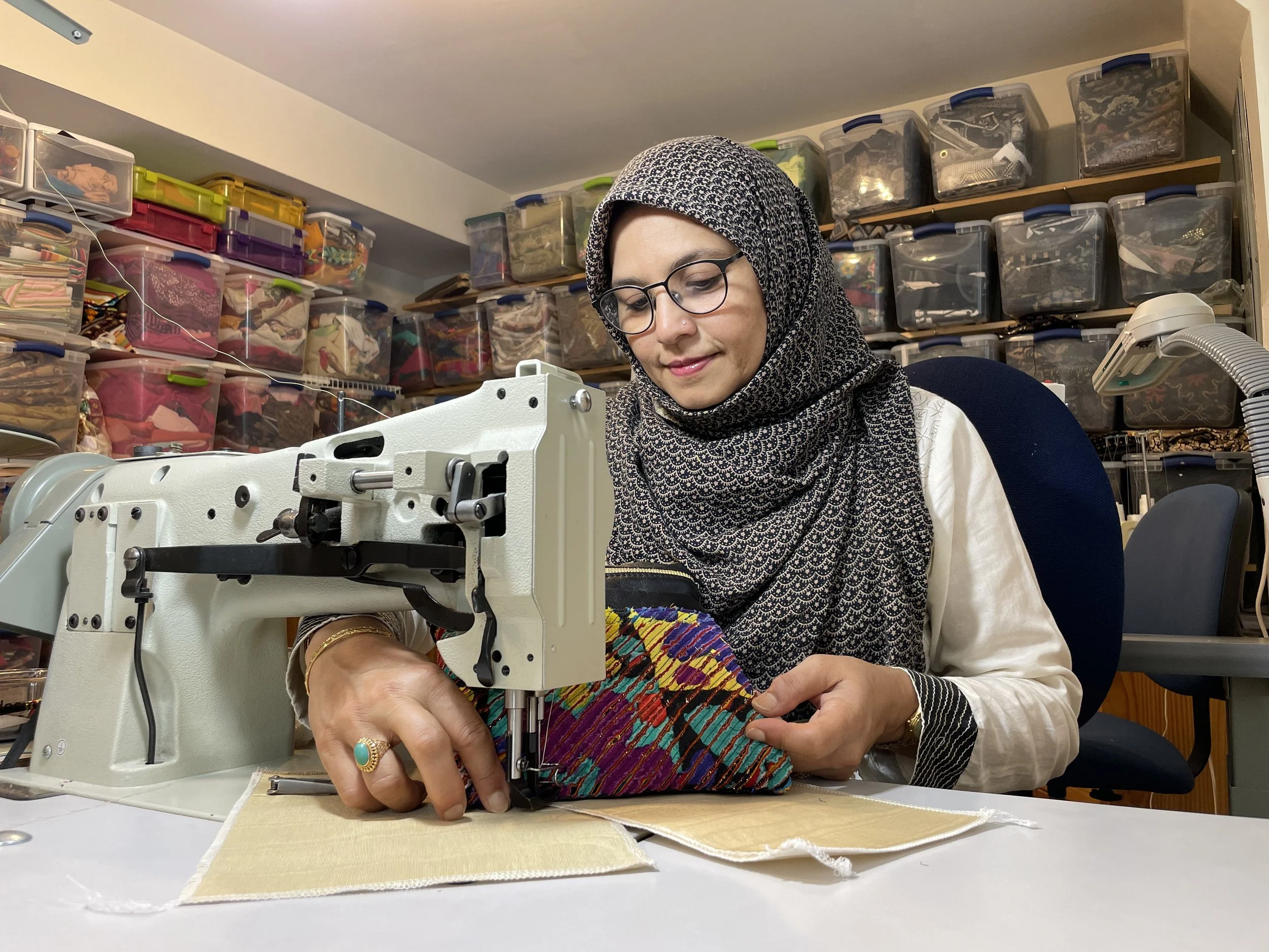 A woman wearing glasses and a patterned headscarf sewing colorful fabric on a sewing machine in a craft room filled with storage bins of fabric and sewing supplies.