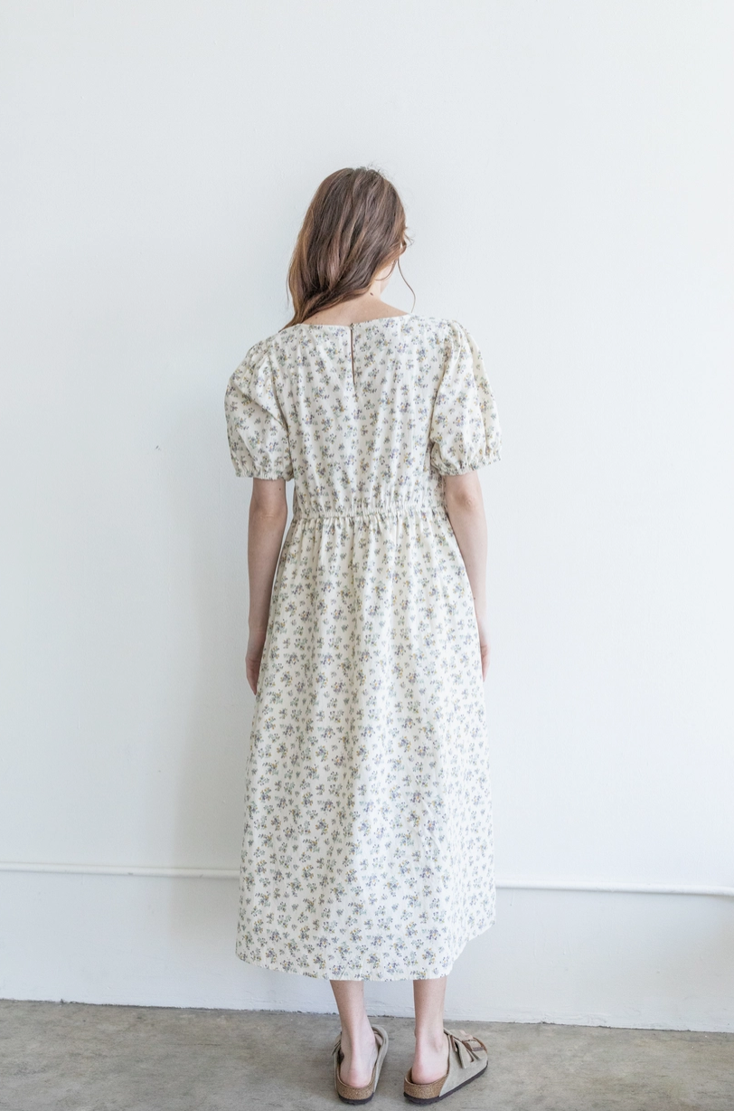 A woman with brown hair wearing a white floral dress and beige sandals, standing against a plain white wall.