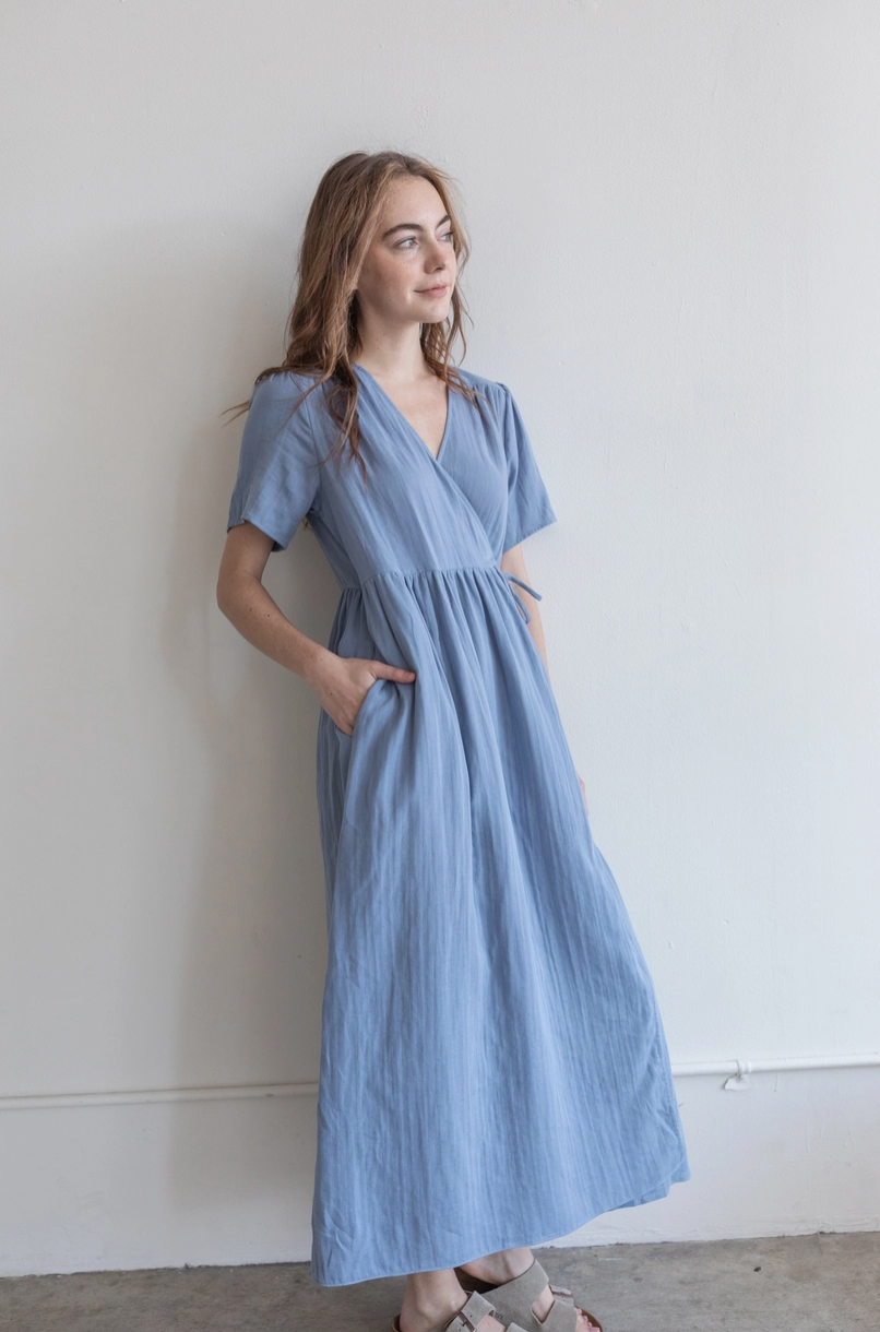 A young woman with light brown hair and fair skin, wearing a blue dress with short sleeves and pockets, stands relaxed against a white wall with her hands in her pockets, gazing to the side.
