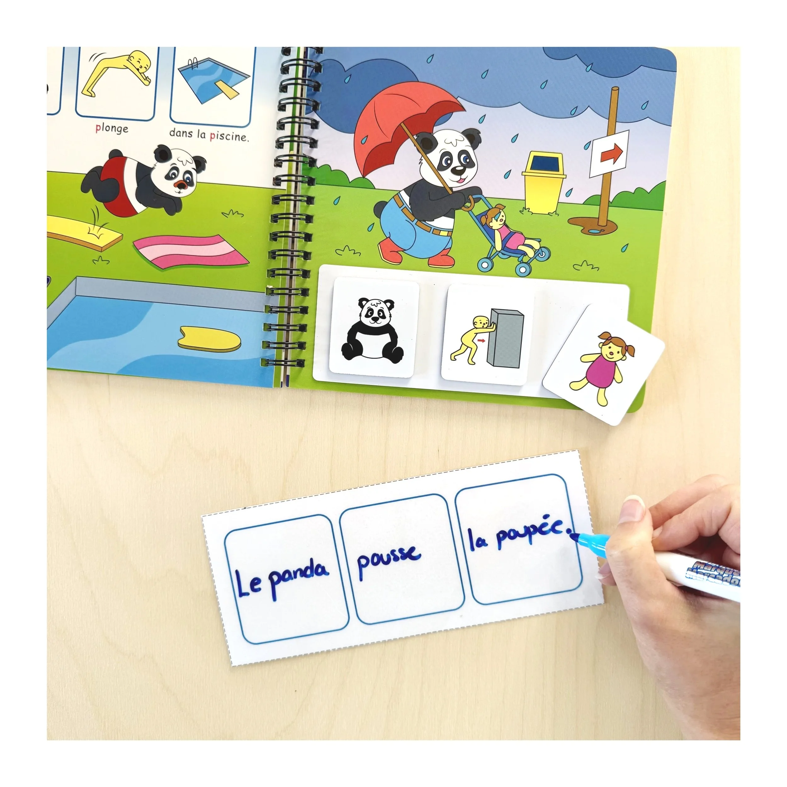 Une page illustrée d'un livre pour enfants avec un panda portant un parapluie, un chien qui saute dans une piscine et des cartes de vocabulaire en français. Une personne écrit à la main des mots sur des étiquettes.