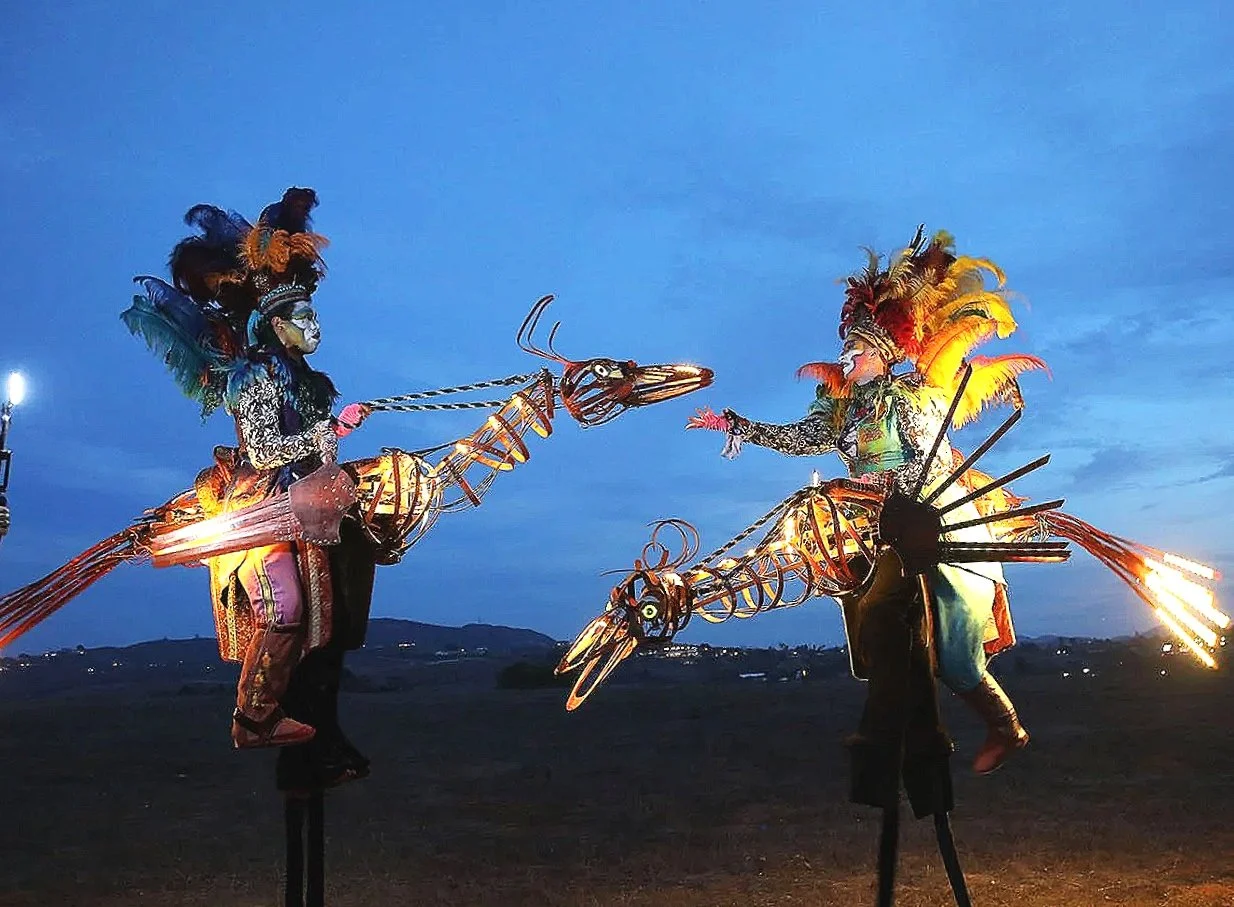 Two stilt performers operating an illuminated dragon stilt puppet during an outdoor night street theatre performance