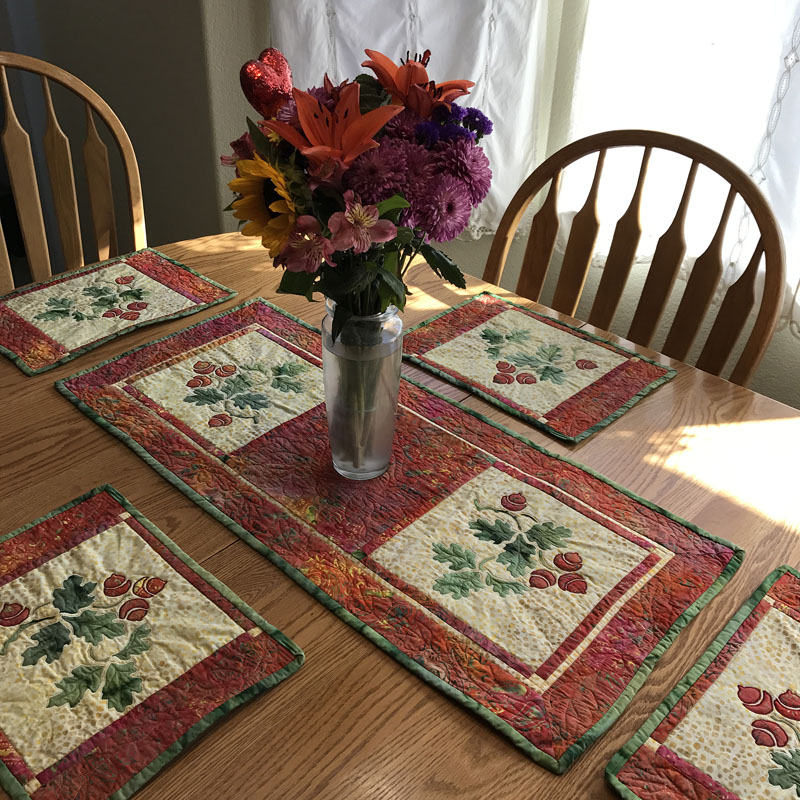Autumn's Promise table decor set. table runner (16" x 35"); placemats ( 10" x 14"). Hand or machine reverse applique with applique, and machine pieced. Designed in fabrics by Timeless Treasures.