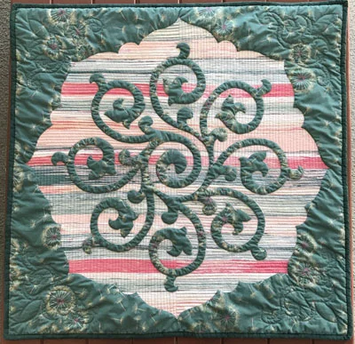 Whirlwind, Medallion VI (27” x 27”). Hand reverse applique, machine quilted. with a double bat of wool on top of cotton. The piece would benefit from added quilting in the surrounding top fabric. Modernesque fabrics by Art Gallery with a modernesque style of machine quilting.