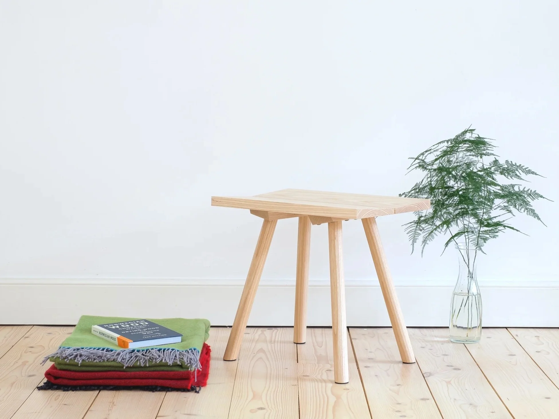 a small wooden contemporary stool next to some textiles on a wooden floor