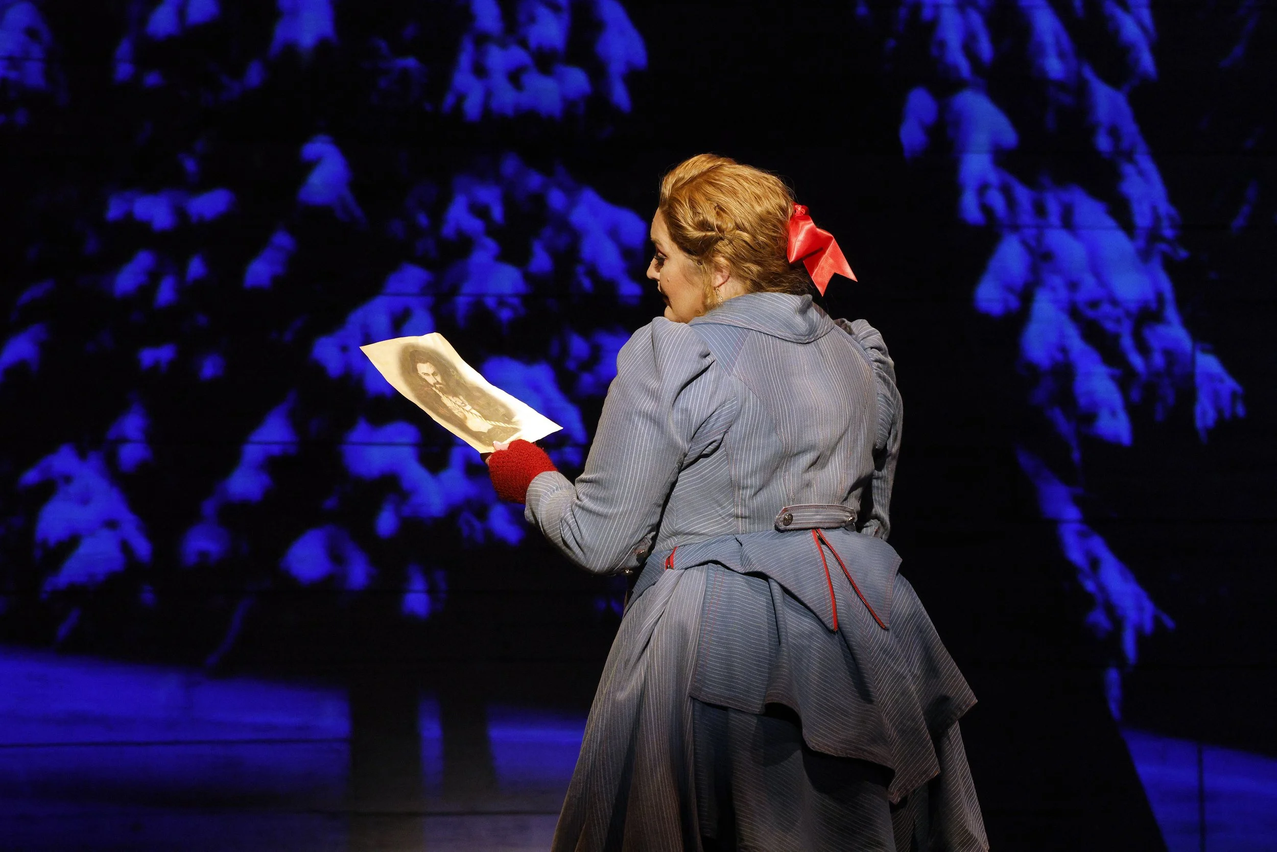 Julie Adams as Senta stands with red hair tied with a red bow, wearing a gray dress, holding the potrait of the Dutchman, standing in front of a dark background with blue-lit trees.