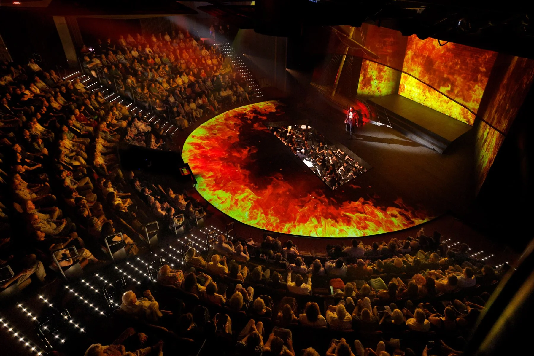 A scene from The Flying Dutchman at Des Moines Metro Opera. An auditorium with a circular stage in the center, displaying fiery visuals on the stage and background screens, filled with an audience watching a live performance or presentation.