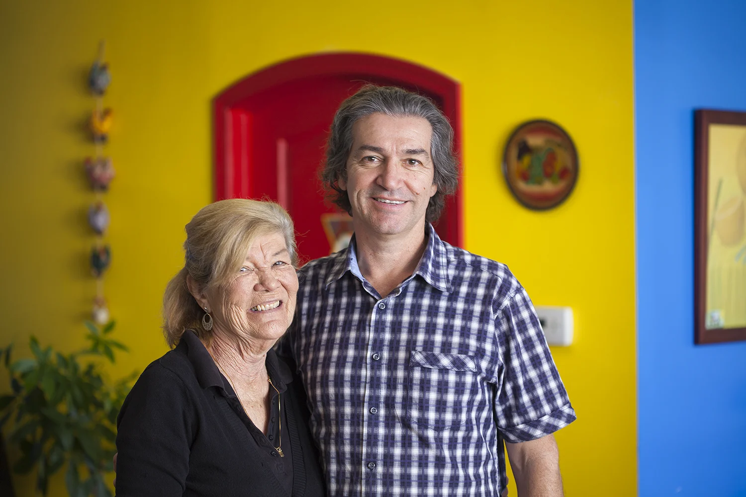 An elderly woman and a middle-aged man smiling and standing together indoors.