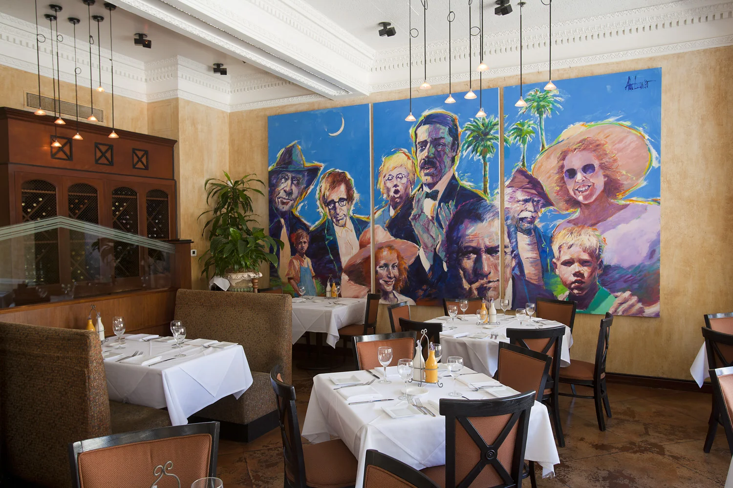 A restaurant interior with tables set with white tablecloths, wine glasses, and utensils. There is a large colorful mural on the wall depicting various people, including a man in a hat, a woman with curly hair, and children, with palm trees and a moo