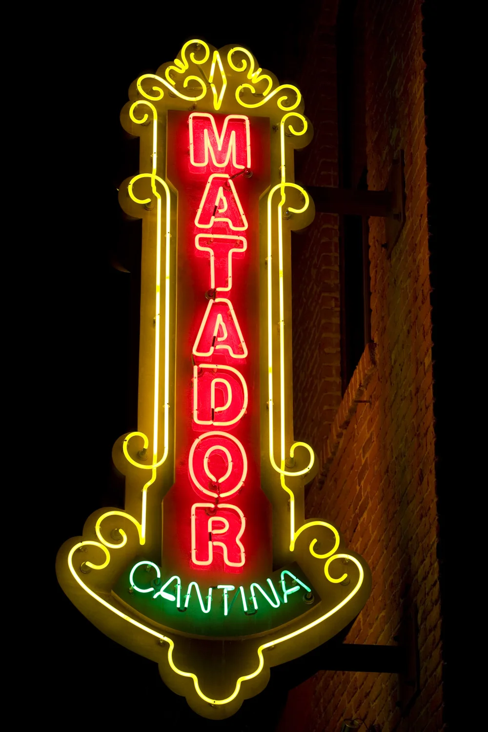 Neon sign with the words "Matador Cantina" in red and yellow lights, mounted on a brick wall at night.