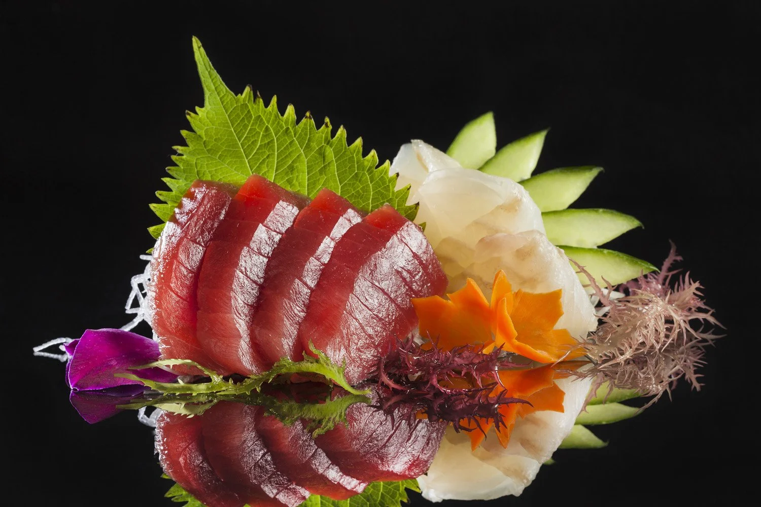 Sashimi platter with slices of raw fish, green leaves, cucumber slices, and colorful edible flowers on a reflective black surface.