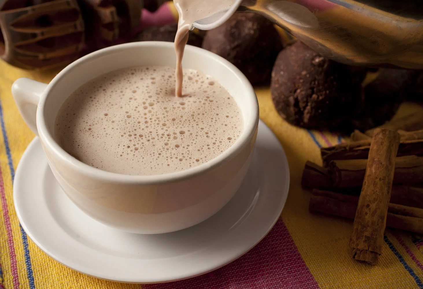 A white cup of hot chocolate with frothy milk, on a saucer on a colorful tablecloth, with chocolate, cinnamon sticks, and spices in the background.