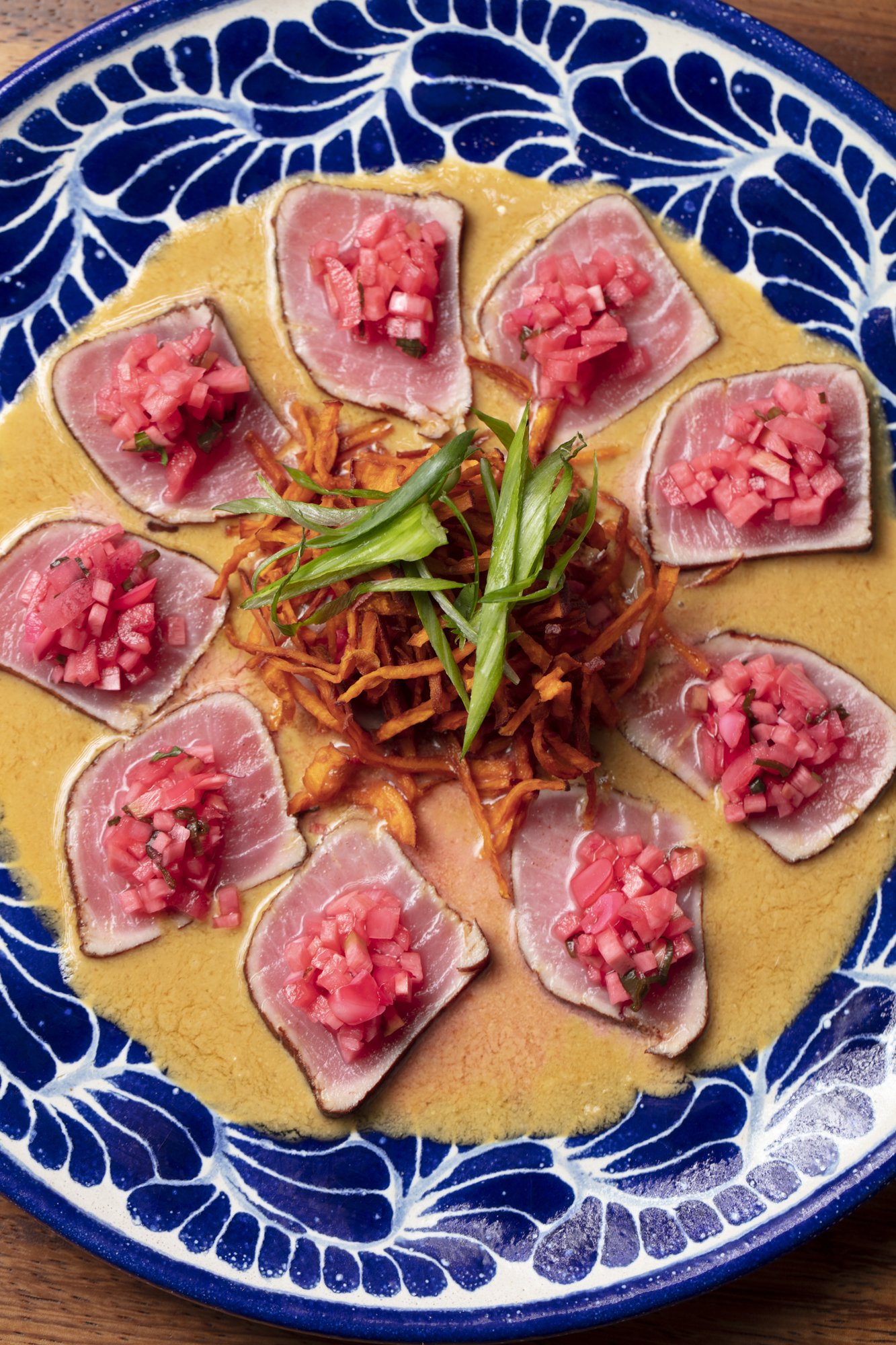 Sliced raw tuna or fish topped with diced pink pickled vegetables, arranged on a yellow sauce or puree, garnished with green onion strips, all served on a blue and white patterned plate.