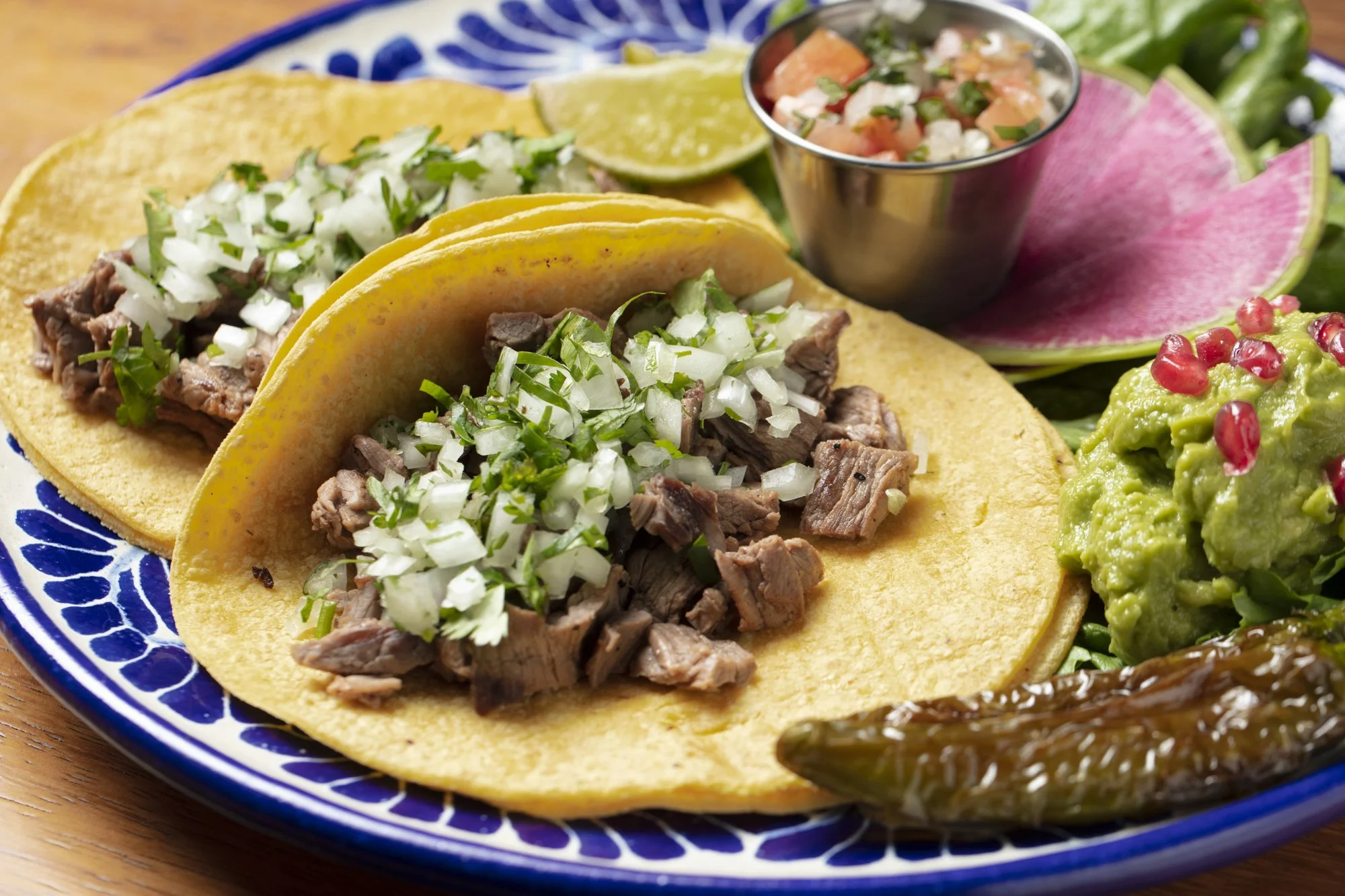 Two beef tacos with chopped onions and cilantro on yellow corn tortillas, served with lime wedges, guacamole topped with pomegranate seeds, roasted jalapeño, and a side of pico de gallo on a colorful blue and white plate.