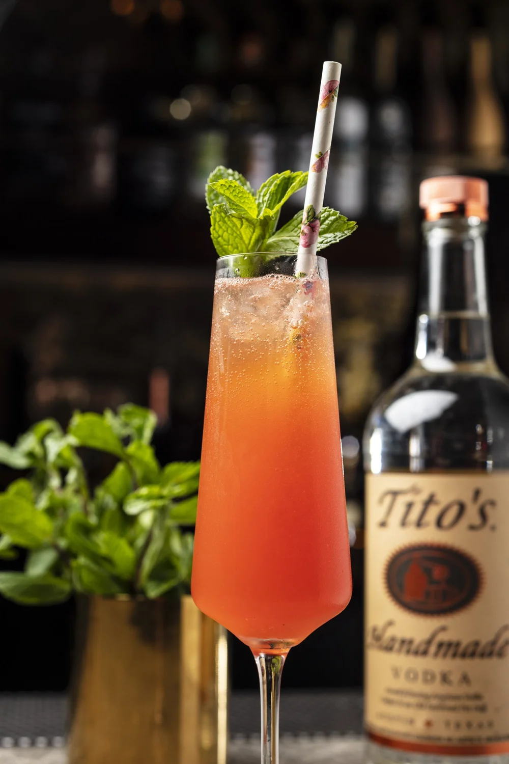 A pink cocktail in a tall champagne flute garnished with fresh mint leaves and a decorative paper straw.
