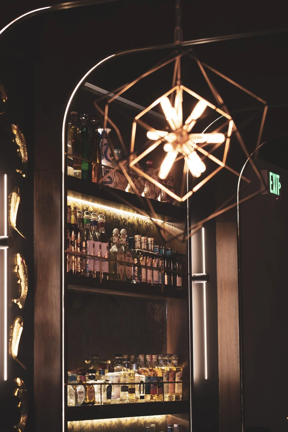 Bar shelf with liquor bottles under a modern geometric hanging light fixture in a dimly lit lounge or bar area