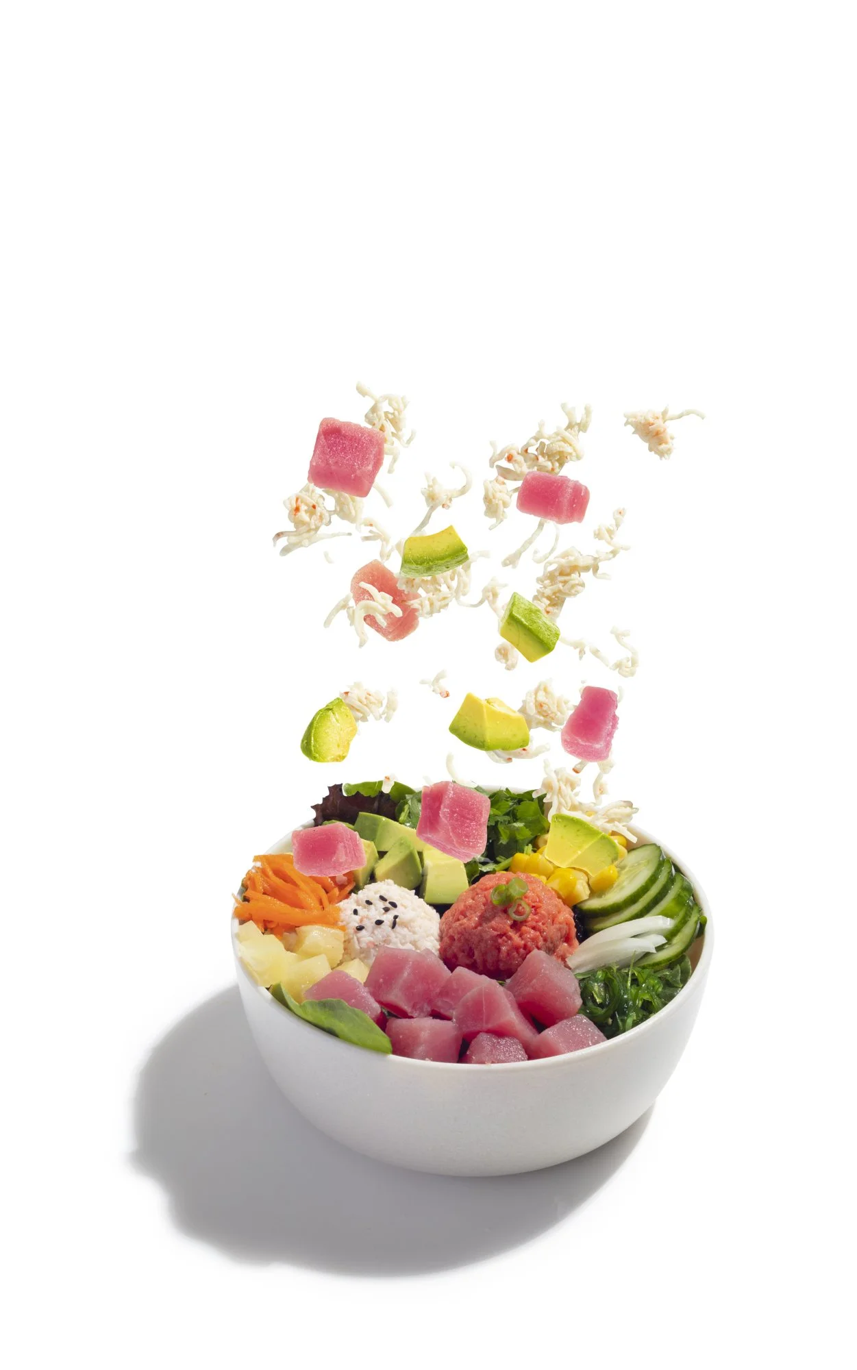 A bowl containing assorted fresh vegetables and tuna, with cubes of watermelon and other ingredients, with some toppings falling into the bowl.